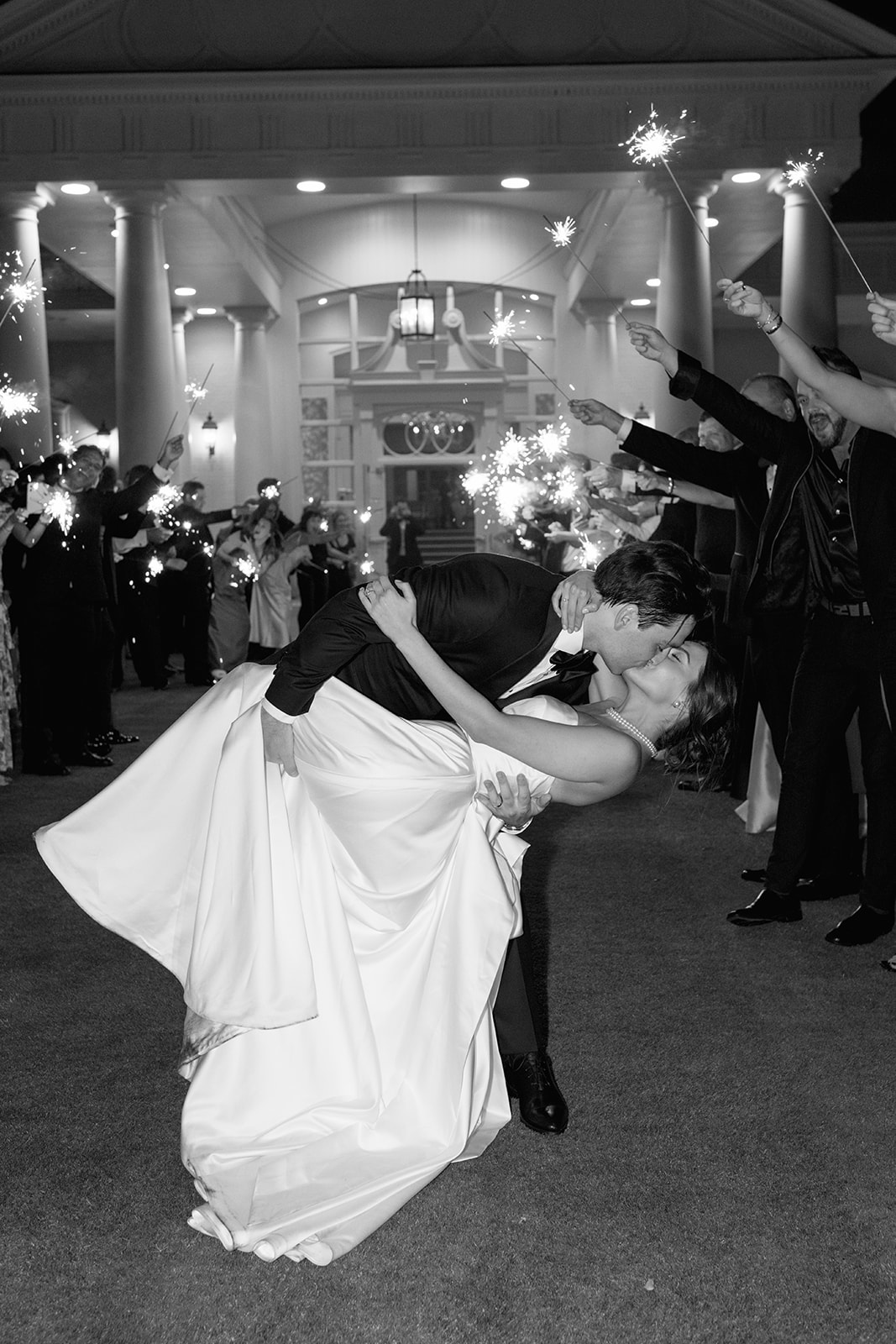 Black-and-white photo of groom dipping bride for a kiss during sparkler exit.