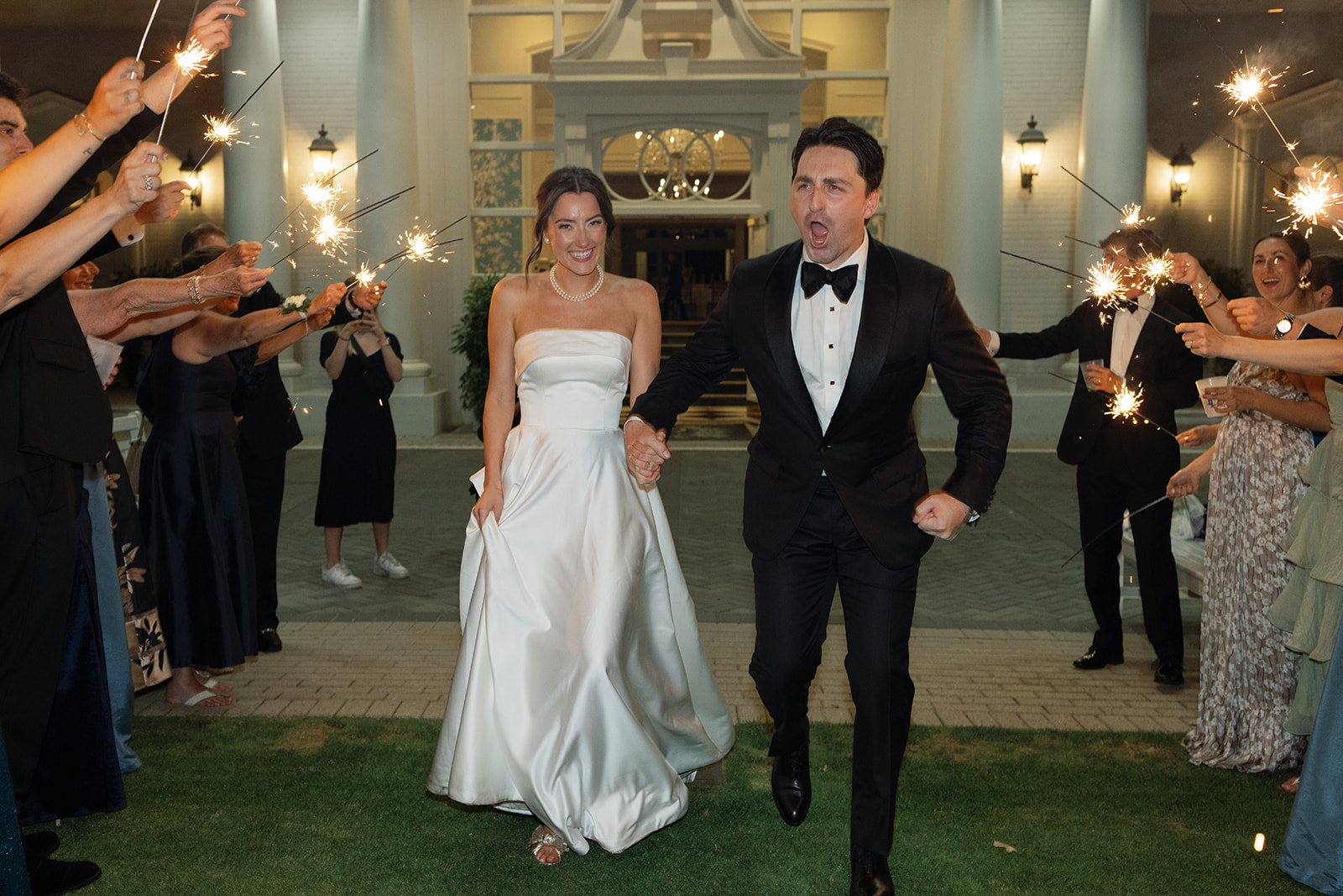 Bride and groom run out of the venue holding hands, guests waving sparklers around them.