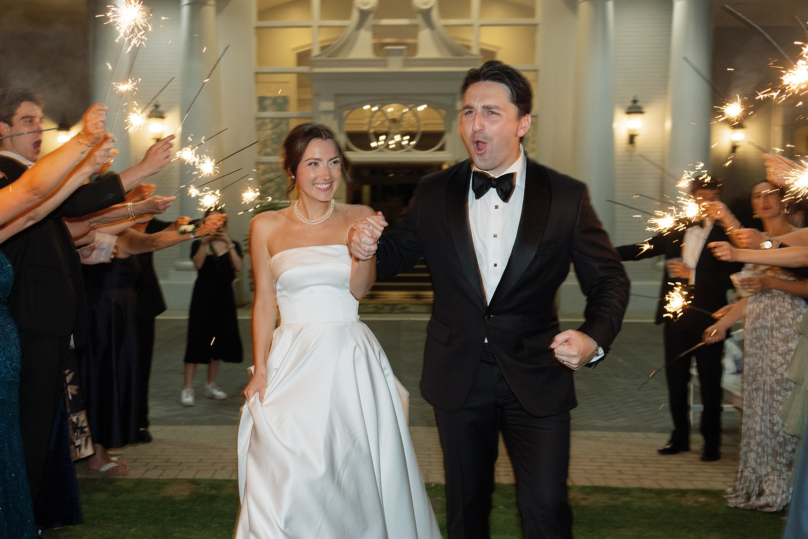Bride and groom run out of the venue holding hands, guests waving sparklers around them.