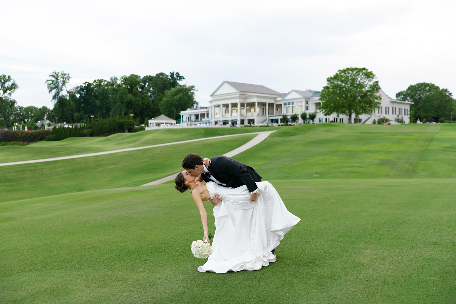 Groom dips bride into a kiss on the green lawn at Myers Park Country Club with the clubhouse in the background.