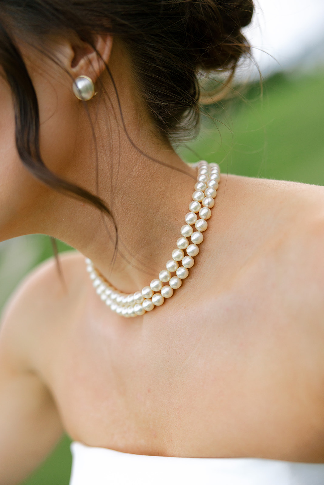 Bride in a strapless gown and pearls smiling softly, holding her bouquet of white roses during pre-ceremony portraits.