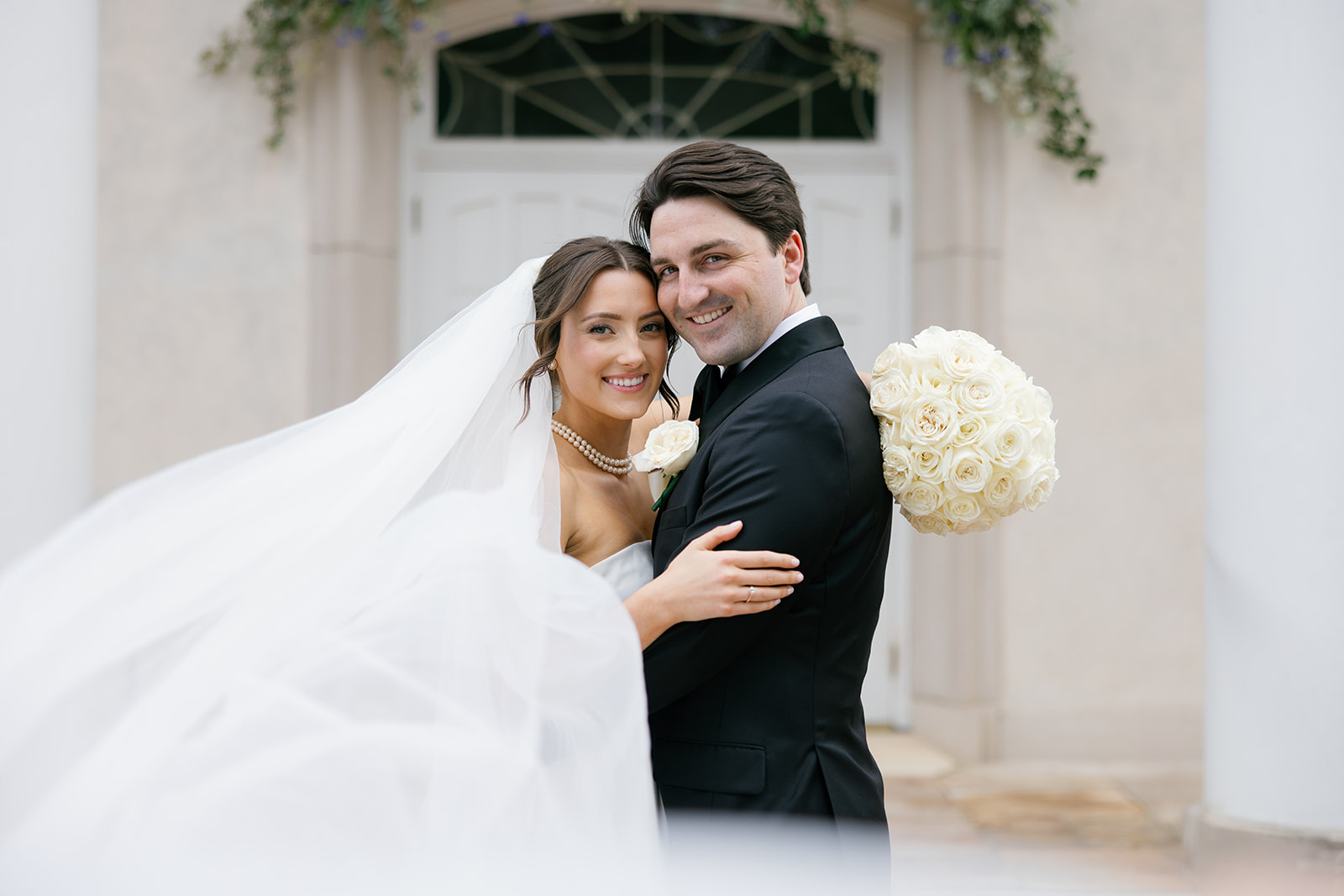 Bride and groom embracing outside the church, smiling at the camera with the bride’s veil flowing around them and a bouquet of white roses behind them.