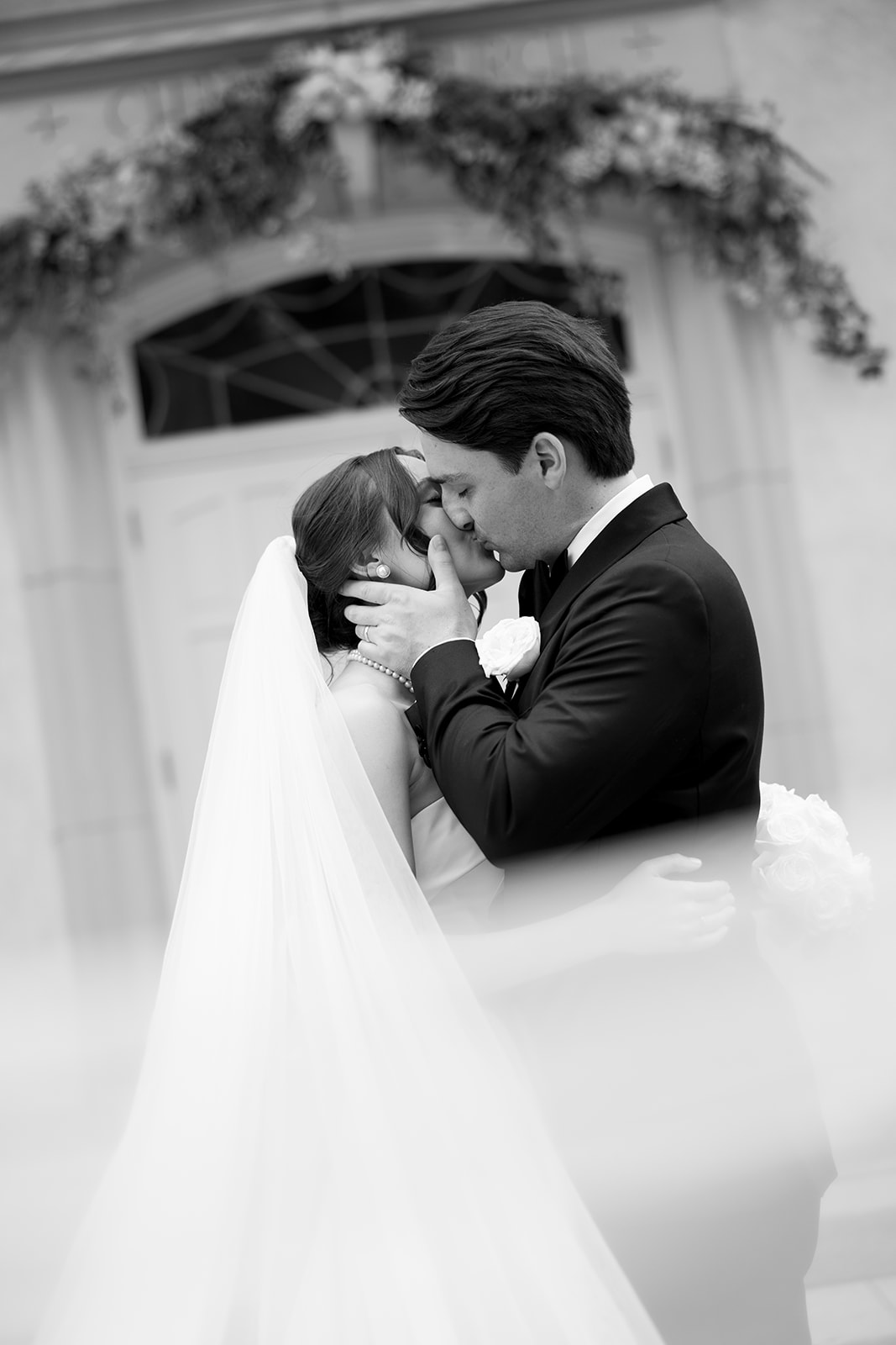 Romantic black and white photo of bride and groom kissing with the bride’s veil softly framing the foreground.