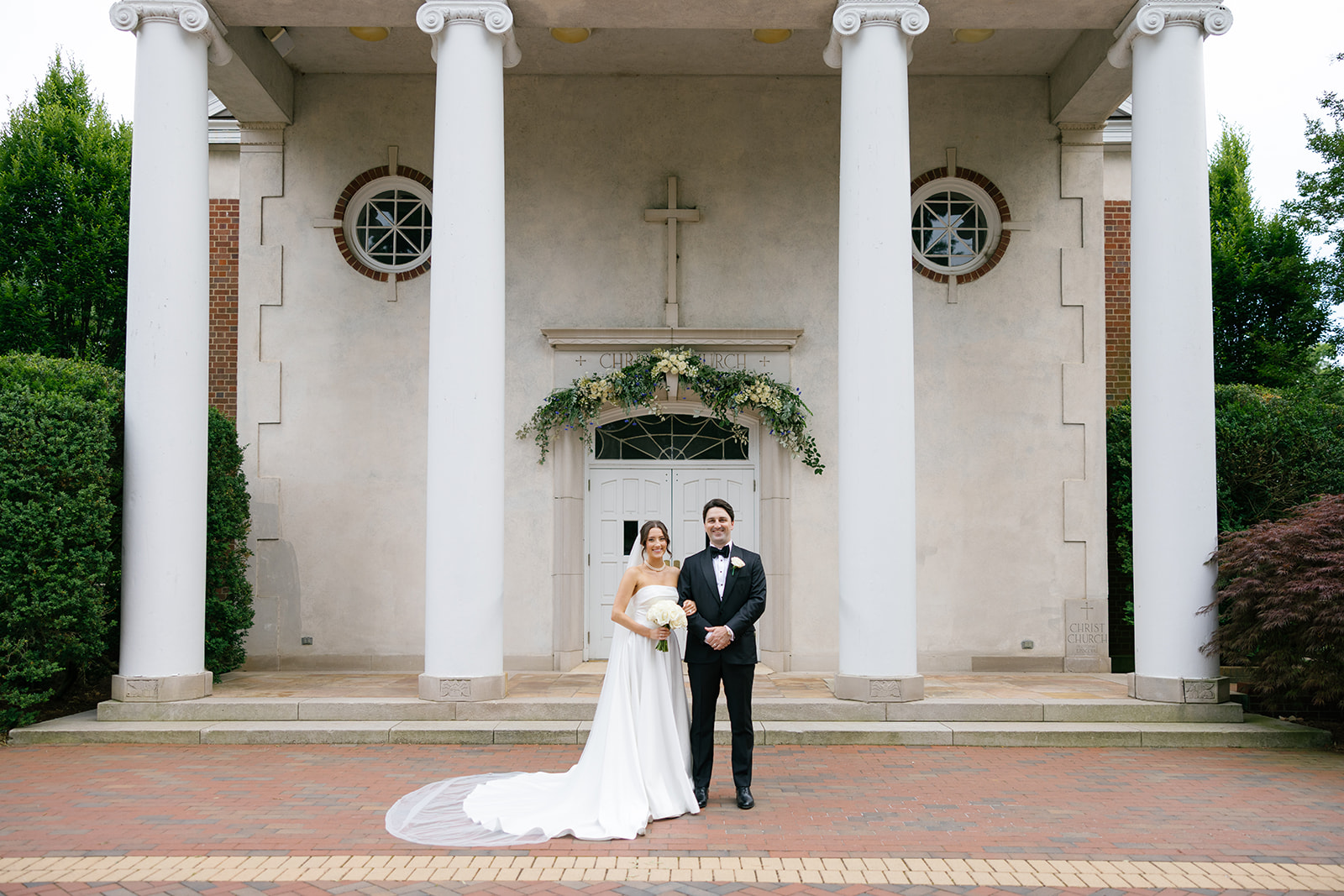 Newlyweds stand in front of the columns at Christ Episcopal Church with floral greenery above the door.