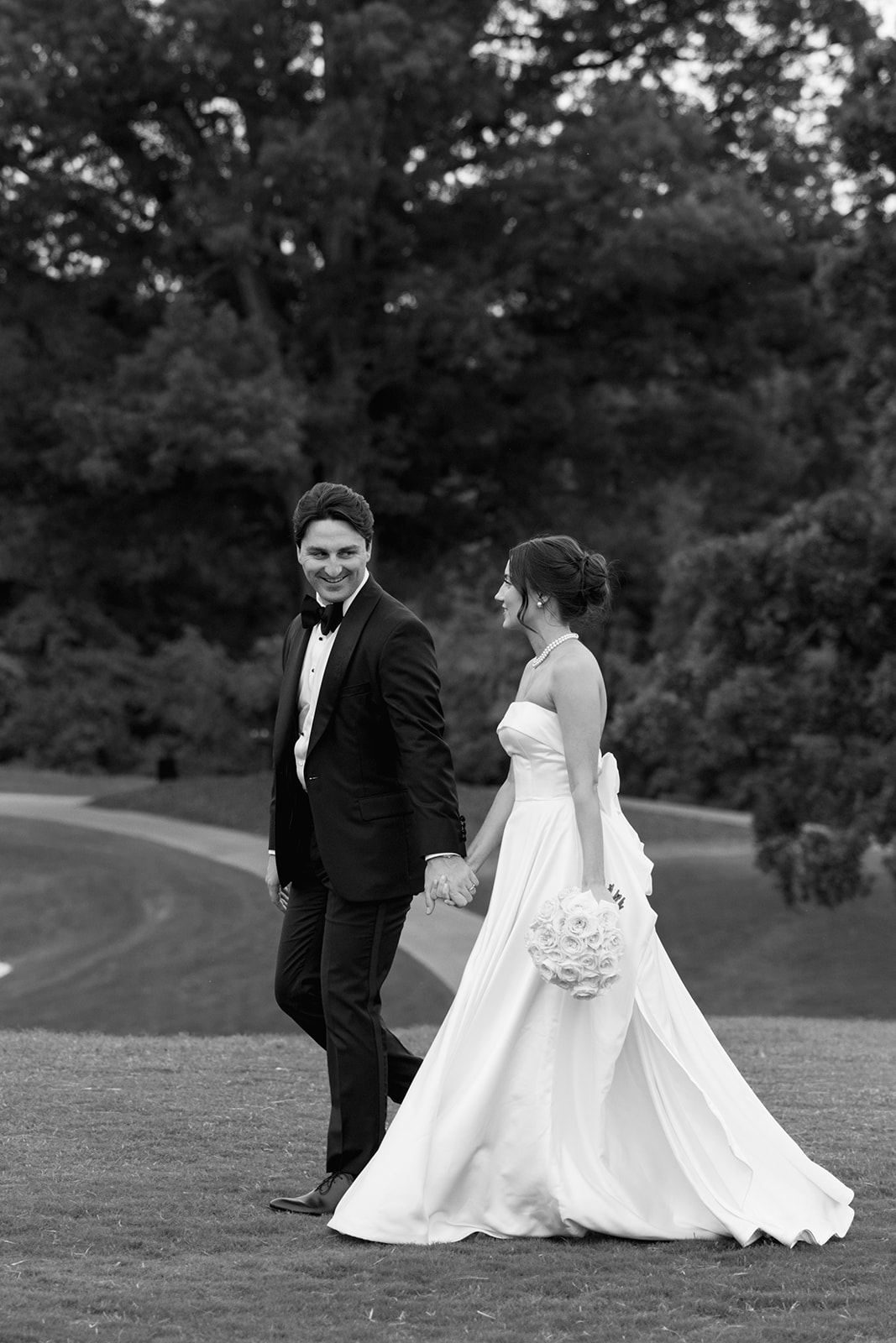 Bride and groom walk hand in hand across the golf course, smiling at each other.