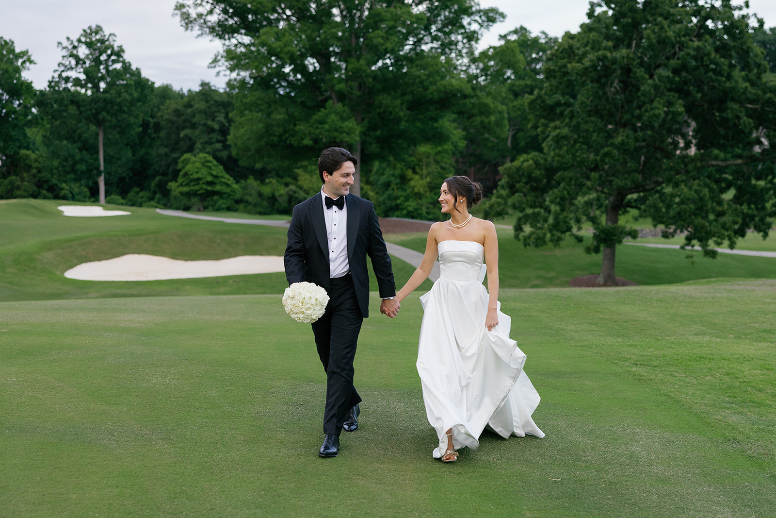 Bride and groom walk hand in hand across the golf course, smiling at each other.