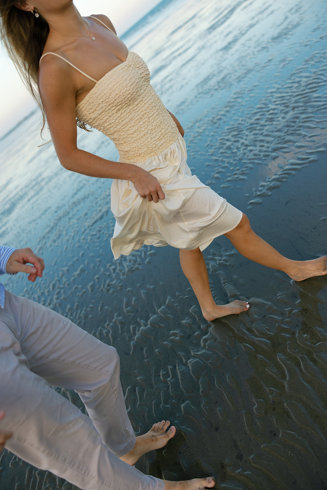 Close-up of a couple walking barefoot along the shoreline during their Charleston SC engagement session, the bride-to-be holding her cream dress above the tide. 