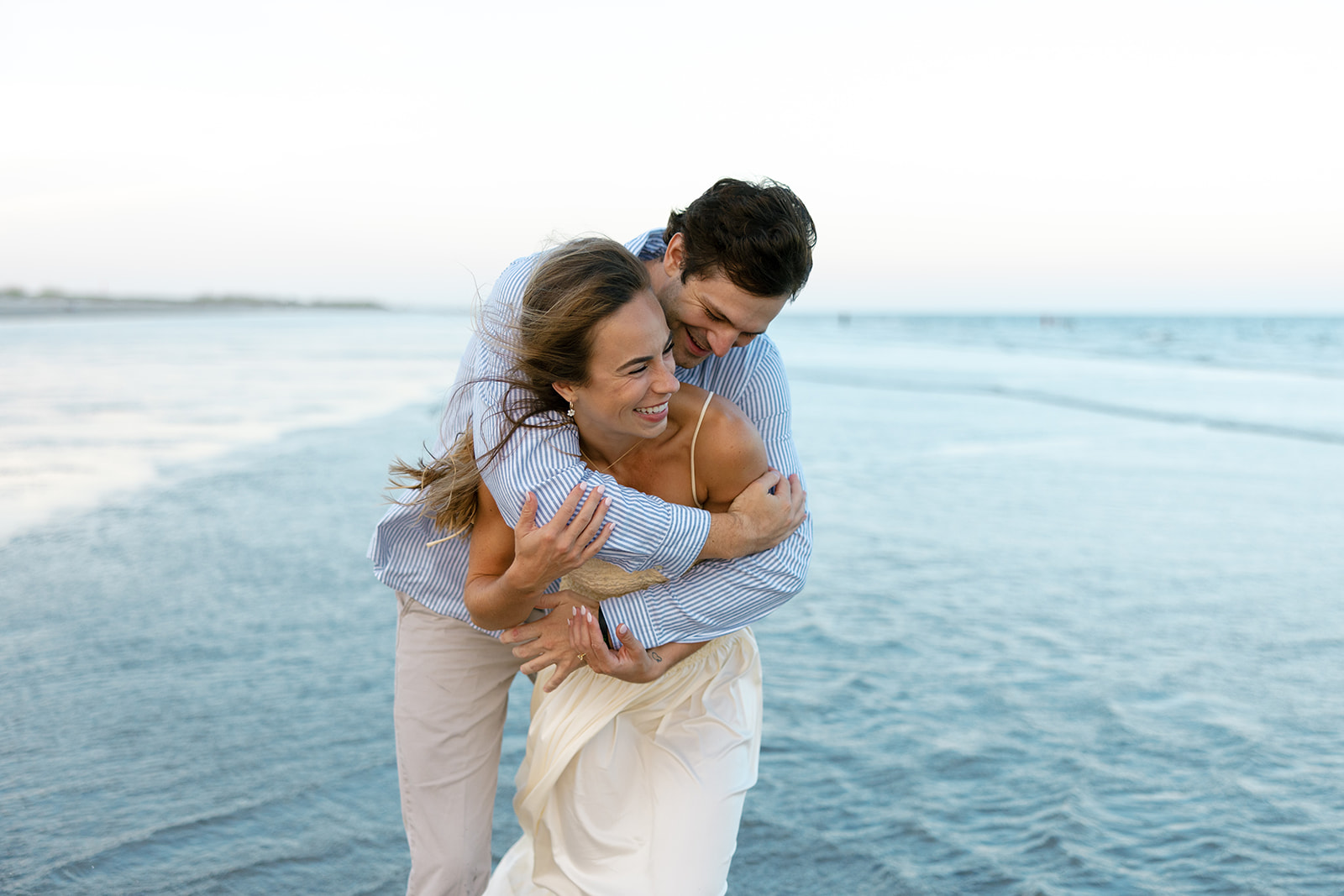 Couple laughing and hugging tightly with ocean waves behind them, a fun and romantic moment captured during their Charleston SC engagement photos.