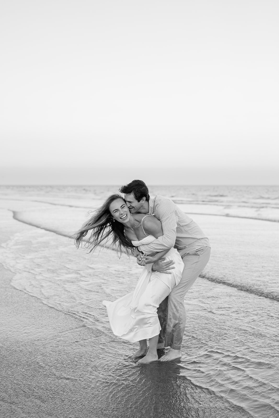 Black and white engagement photo of the couple laughing together as he dips her at the water’s edge on Sullivan’s Island, taken by a Charleston SC engagement photographer.