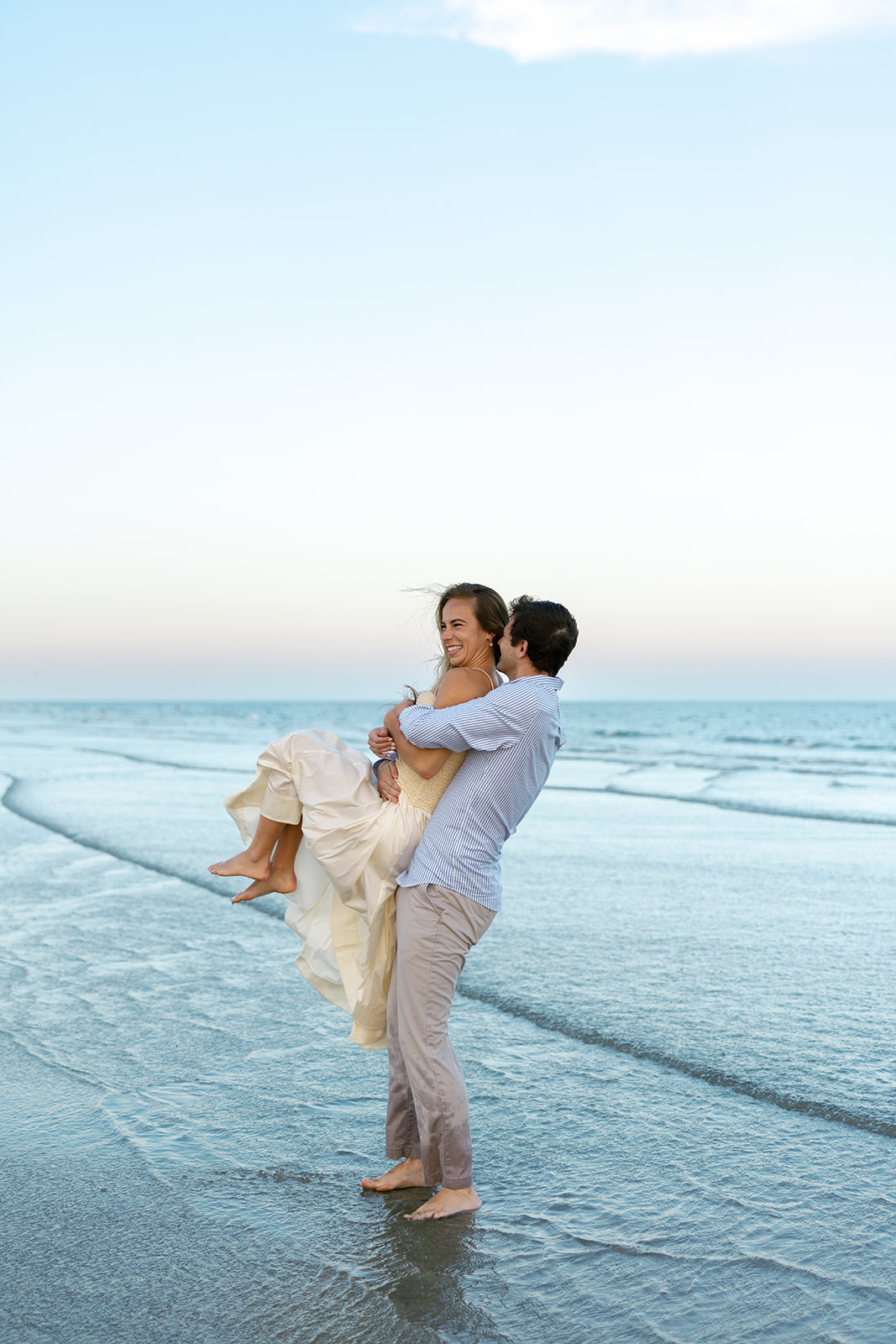 Groom-to-be spinning his fiancée around in his arms as they laugh, standing in the shallow tide.
