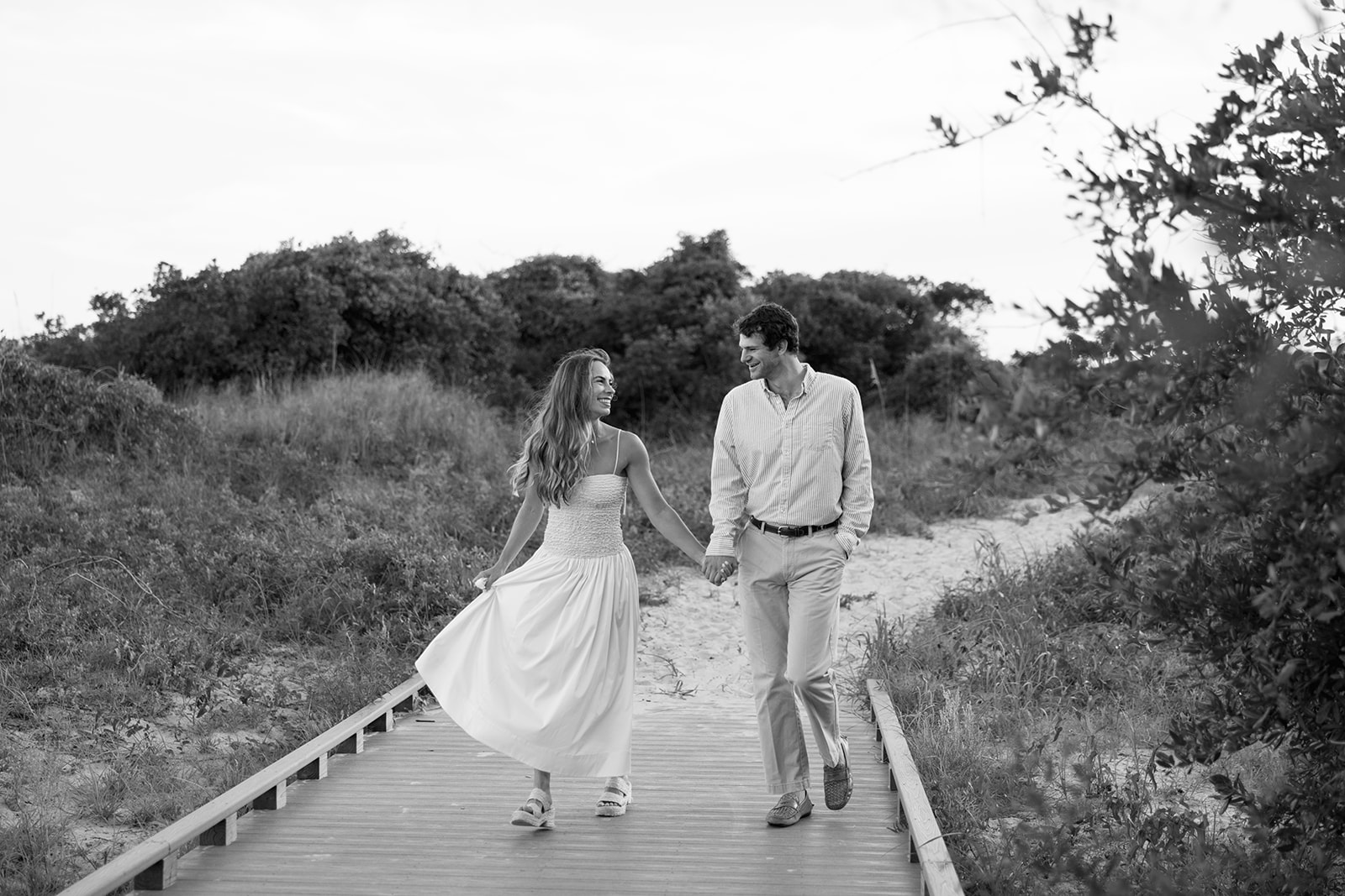 Black and white image of the couple walking hand in hand along the Sullivan’s Island boardwalk, captured by a Charleston SC engagement photographer.