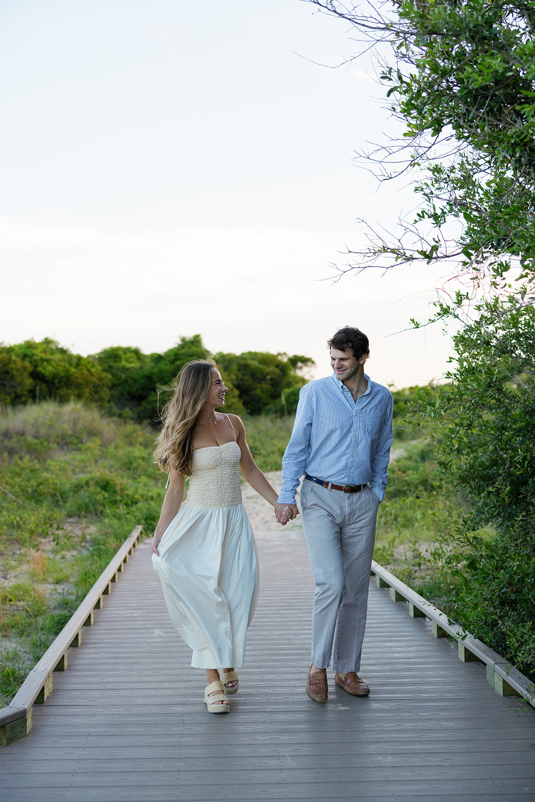 Couple holding hands and walking down a wooden boardwalk surrounded by greenery, captured by a Charleston SC engagement photographer.