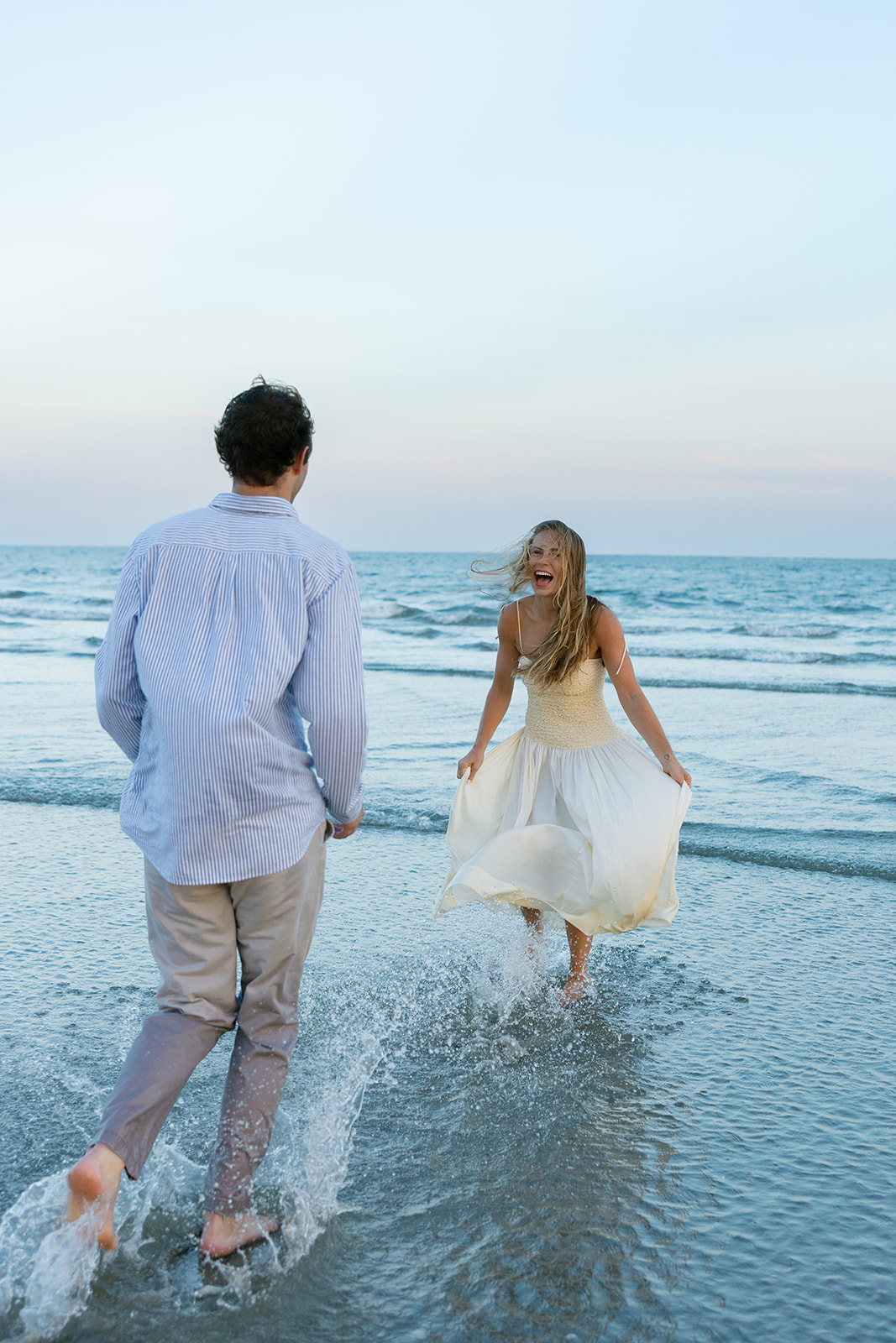 Woman in a cream dress laughing while running toward her partner in a striped shirt across the shoreline.