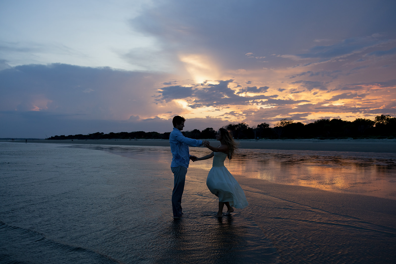 Couple spinning and laughing barefoot in the water at Sullivan’s Island during their Charleston SC engagement session, the vibrant sunset making the perfect backdrop.