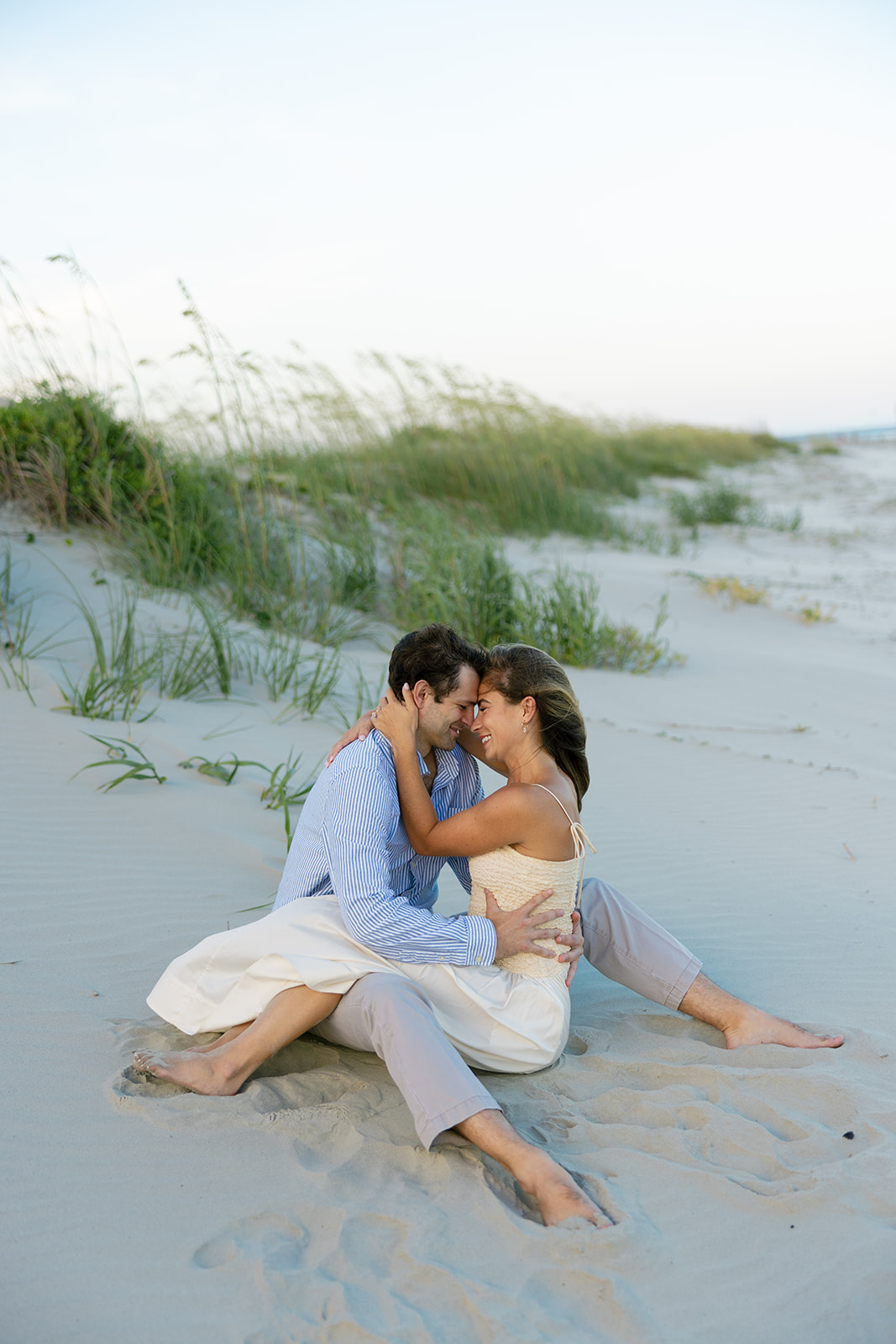 Couple sitting barefoot in the sand dunes at Sullivan’s Island, forehead to forehead, captured by Charleston SC engagement photographer.