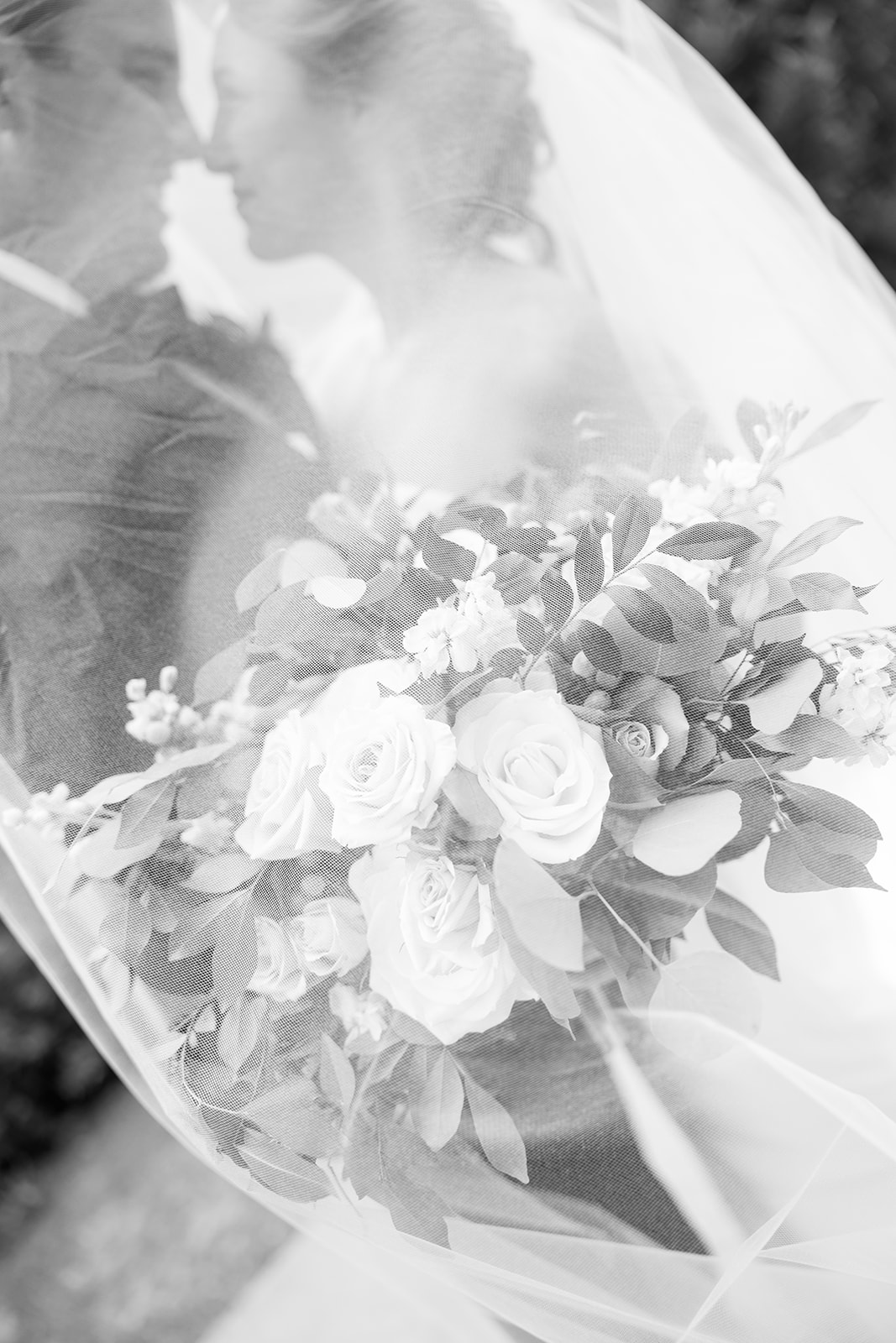 Artistic black and white close-up of the bridal bouquet with the couple blurred behind the veil.