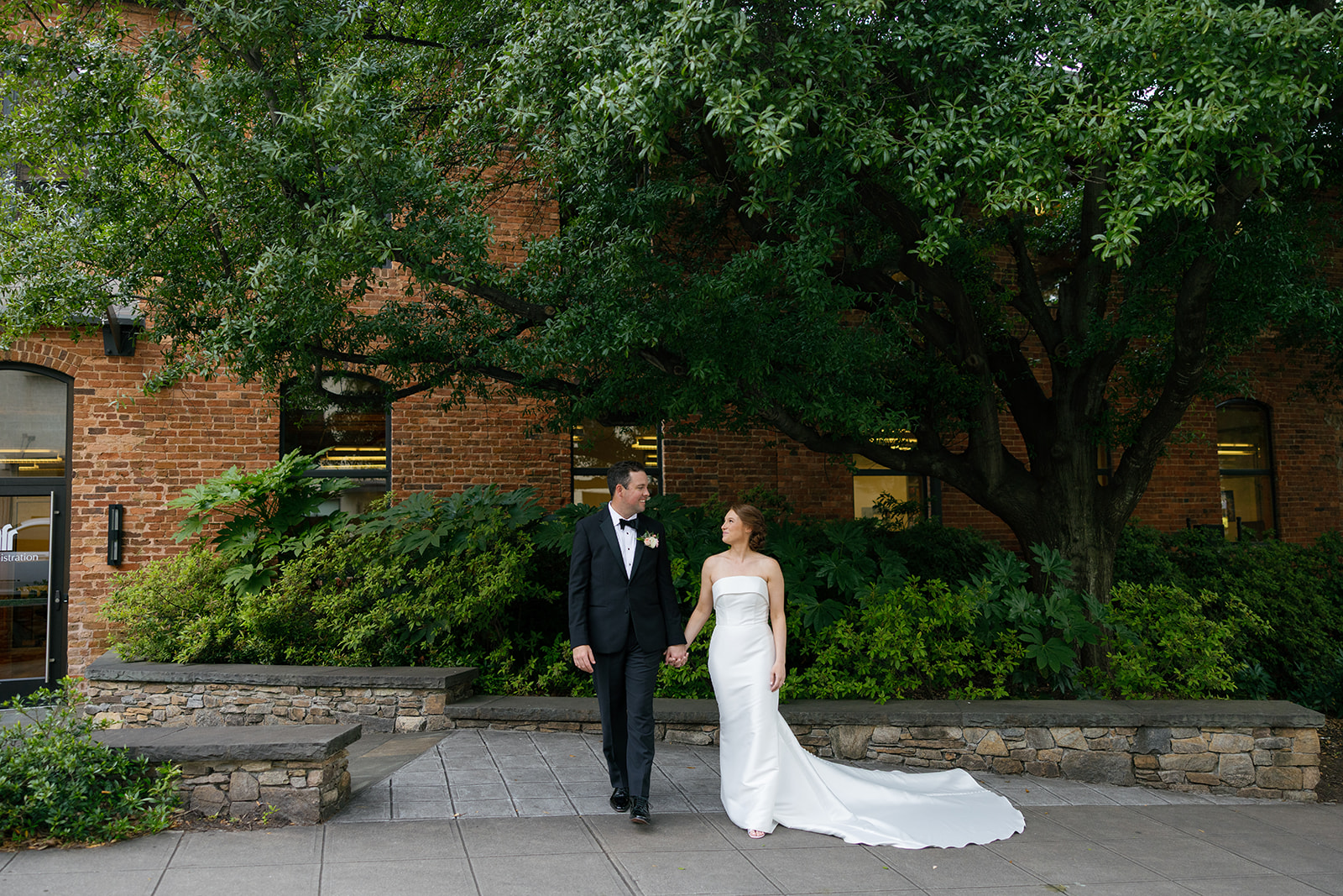Bride and groom walking hand in hand beneath the trees outside Huguenot Loft, smiling at each other.
