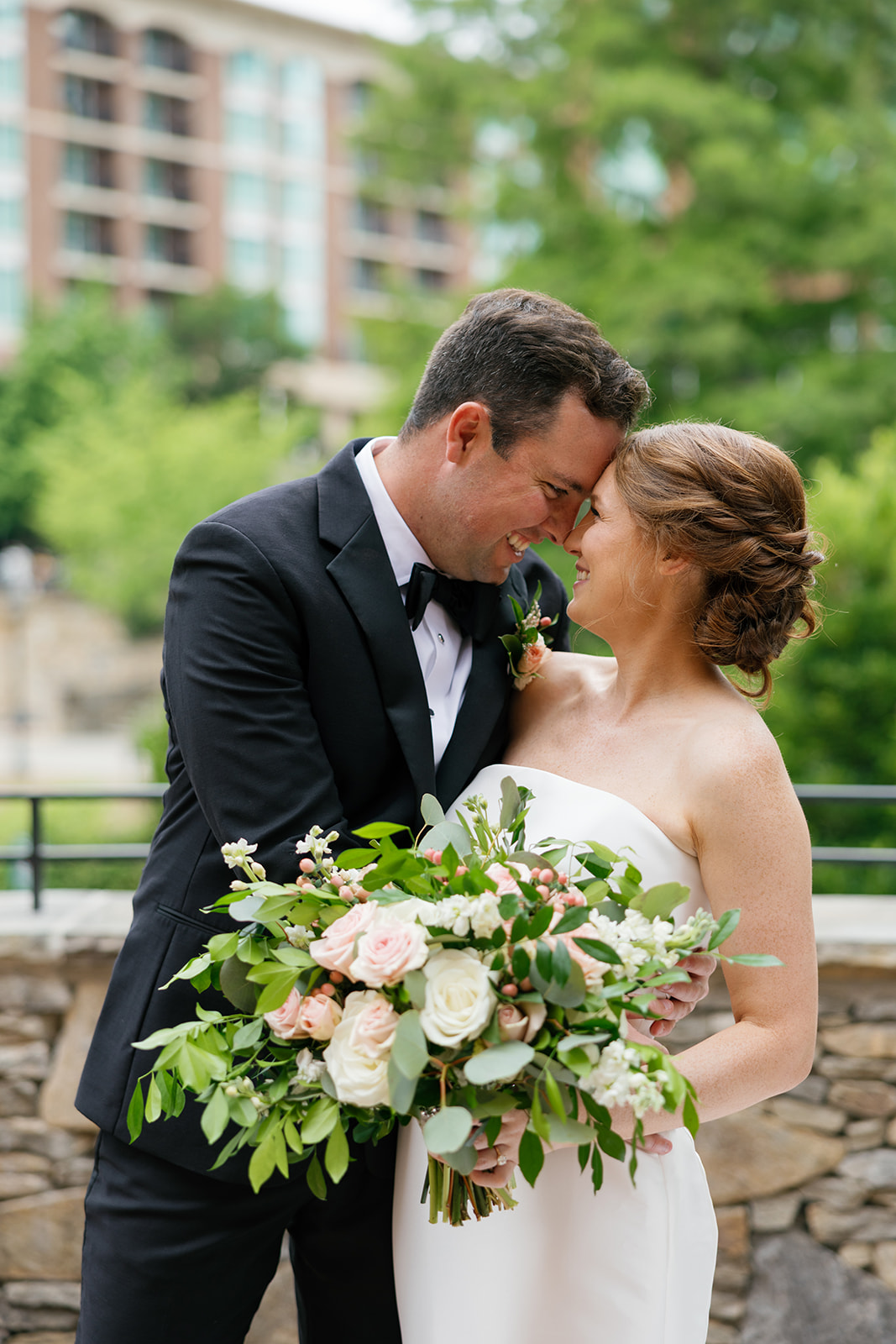 Bride and groom laughing forehead-to-forehead while holding a lush bouquet of blush and white roses outside Huguenot Mill in Greenville, SC.