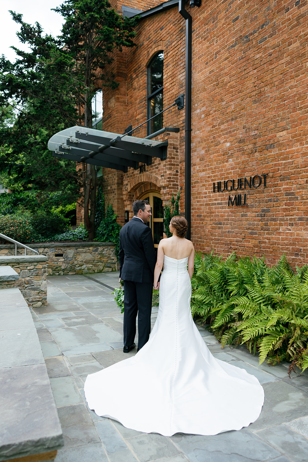 Bride and groom holding hands, standing outside the brick Huguenot Mill with her dress train trailing behind.