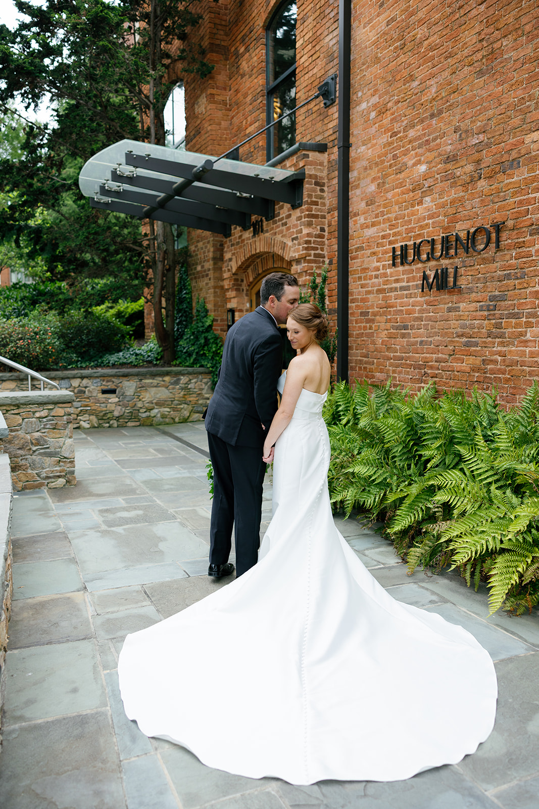 Groom kissing the bride’s forehead as they stand in front of the historic Huguenot Mill brick wall.