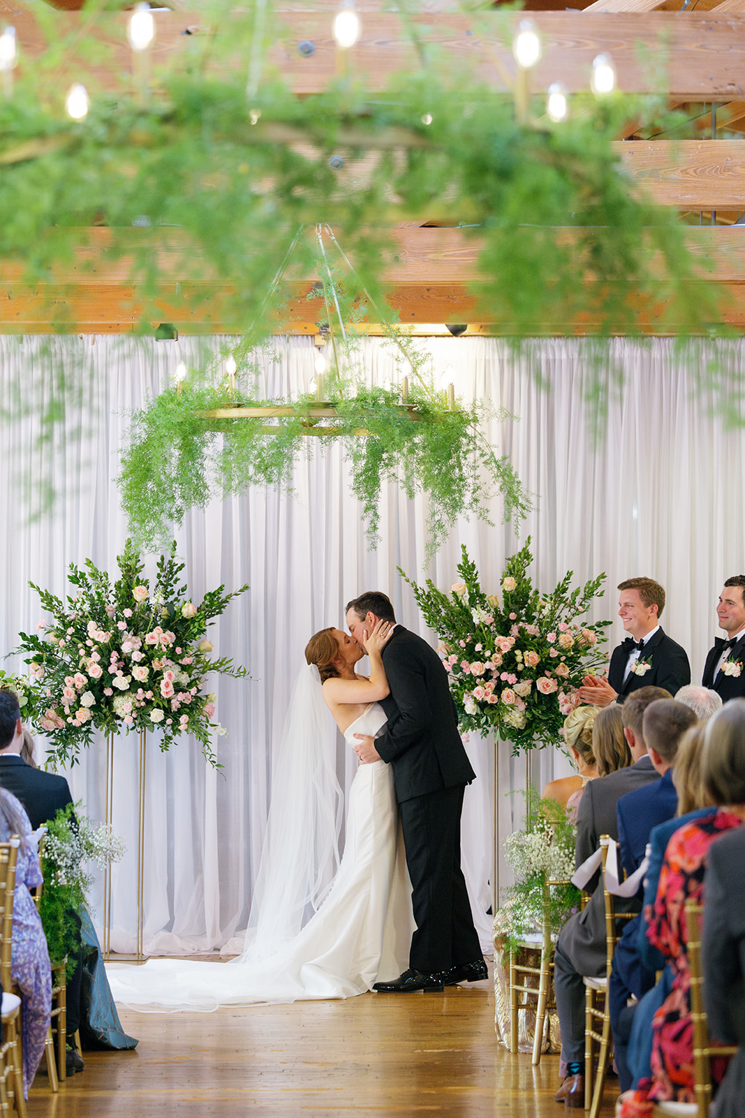 Bride and groom’s first kiss at the altar framed by lush floral arrangements and greenery chandeliers.