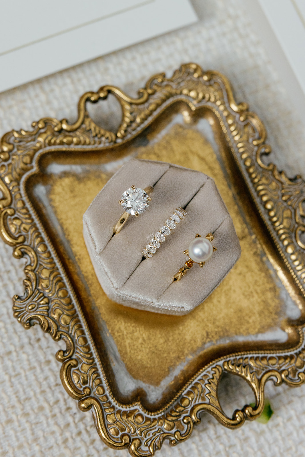 Gold engagement ring, diamond band, and pearl ring displayed in a velvet ring box on an ornate vintage tray.