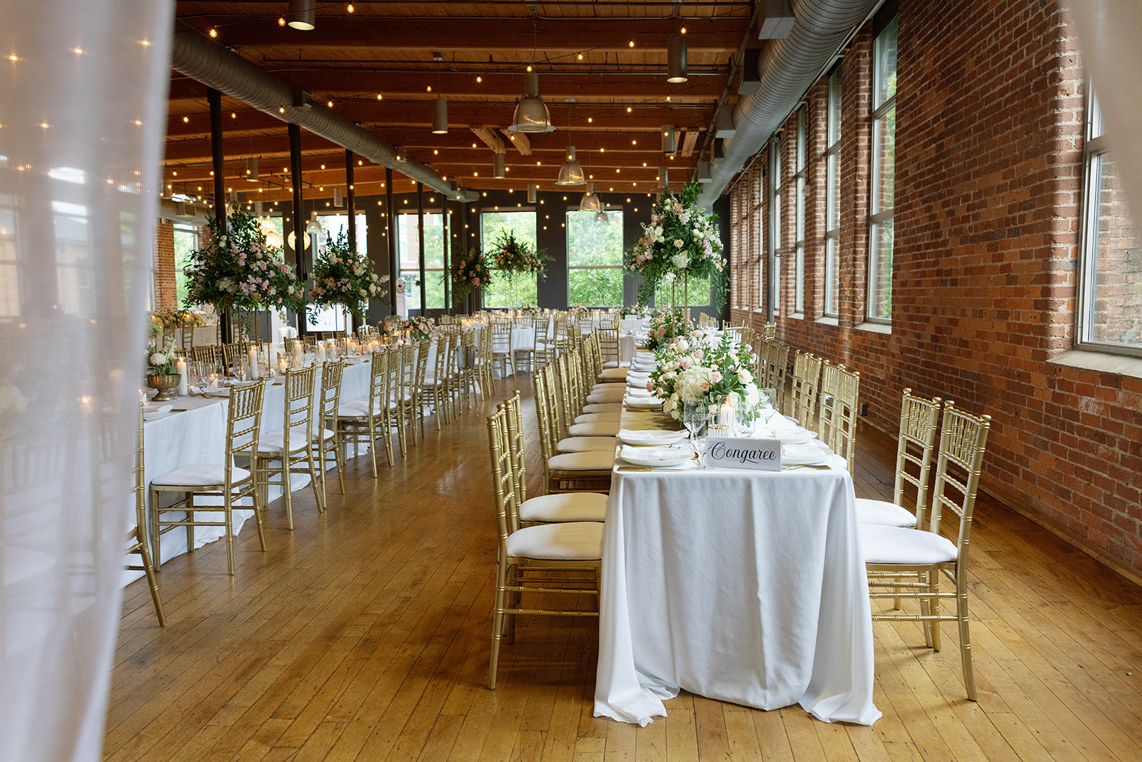 Reception hall at Huguenot Loft and Mill featuring long banquet tables, gold chiavari chairs, and greenery centerpieces in Greenville SC
