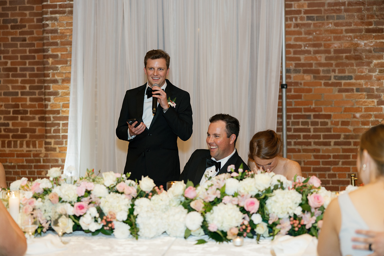 Groomsman delivers a toast during the reception as the groom laughs and the bride covers her face with joy.