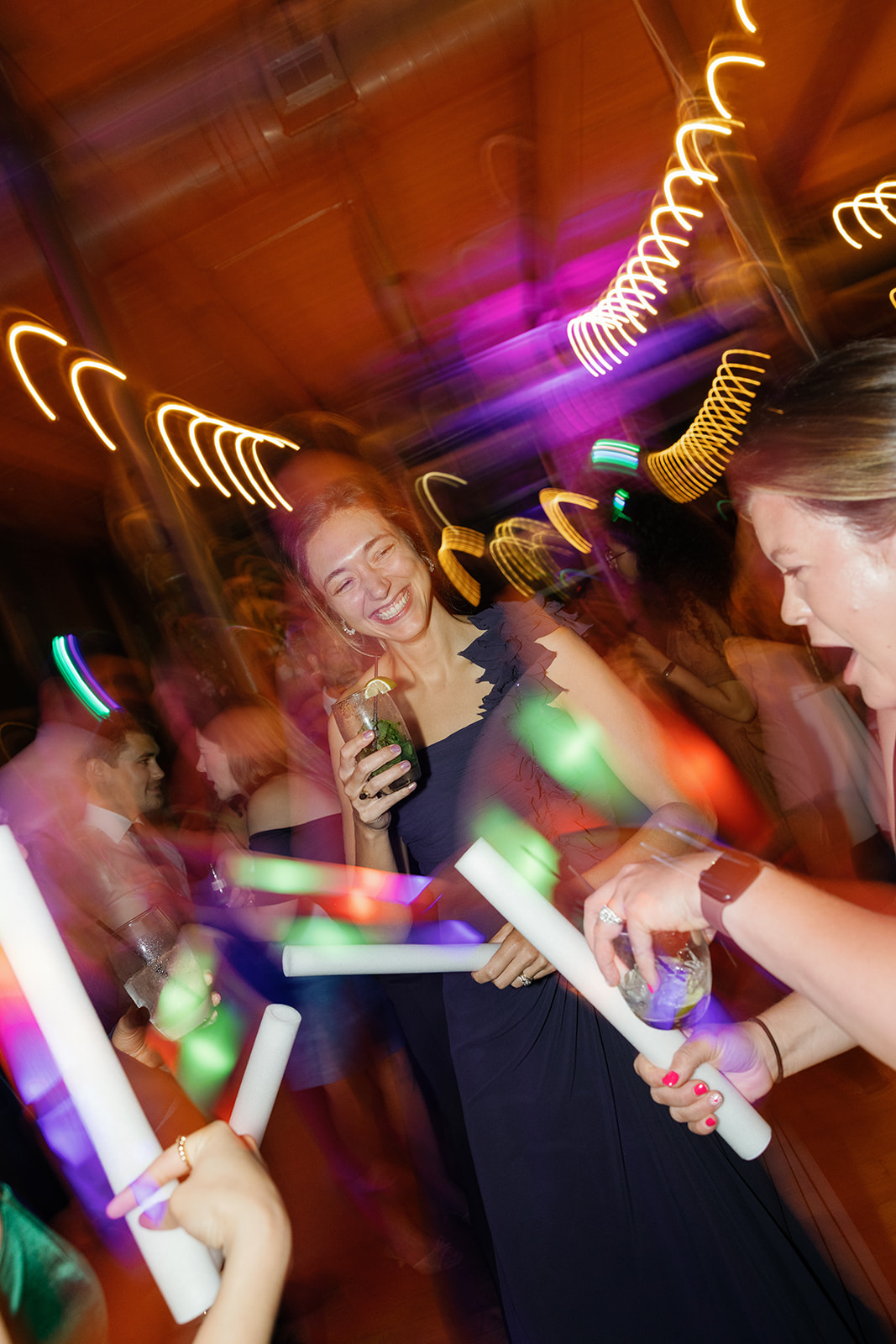 Wedding guest laughing and holding a drink on the dance floor at Huguenot Loft and Mill in Greenville SC, surrounded by glow sticks and brick walls.