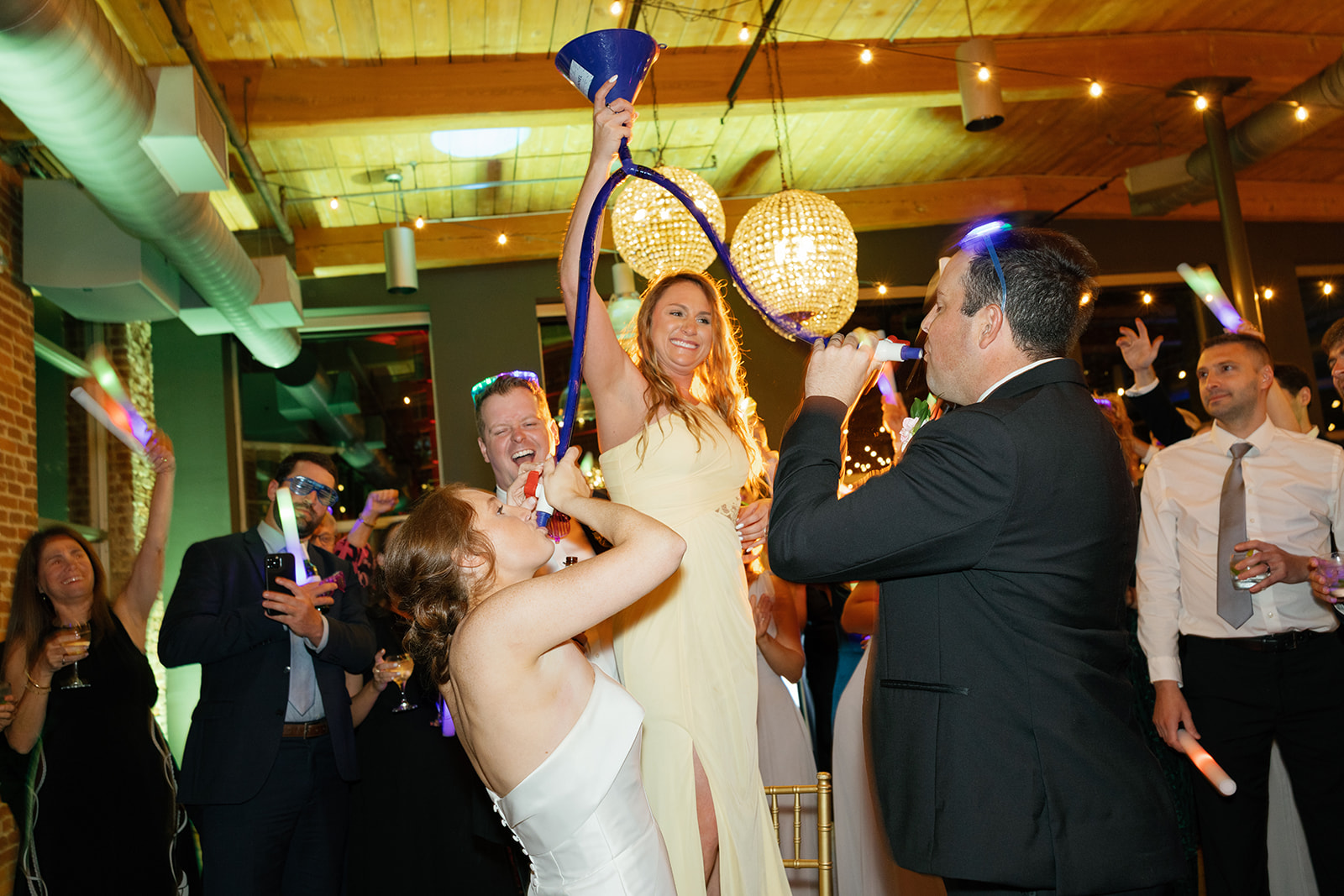 Bridesmaid holds a beer funnel while bride and groom drink, surrounded by cheering guests at Huguenot Loft.