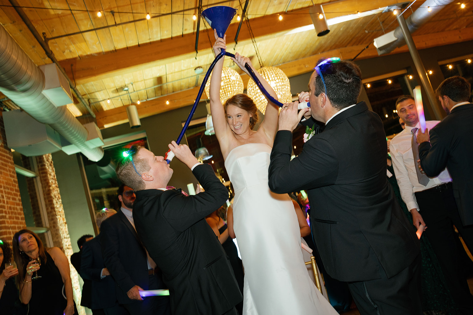 Bride in strapless gown holds a beer funnel while two groomsmen drink, surrounded by cheering guests at Huguenot Loft.
