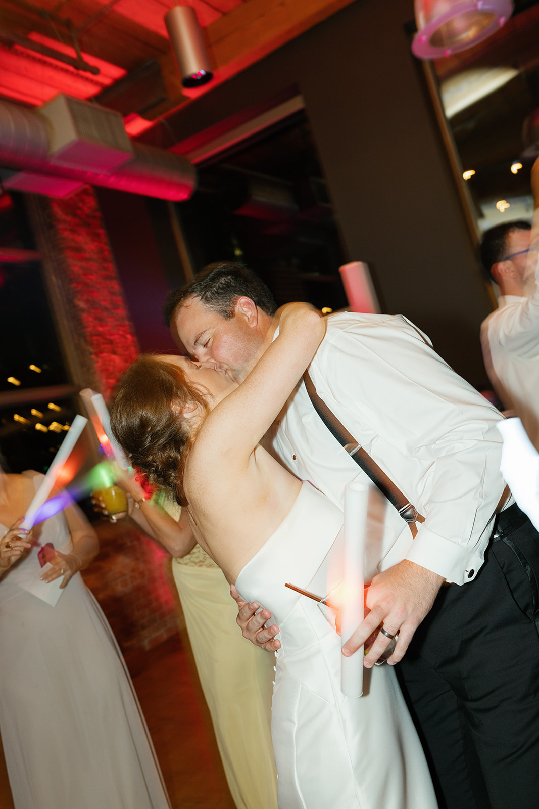 Bride and groom share a kiss on the dance floor at Huguenot Loft surrounded by friends waving glow sticks.