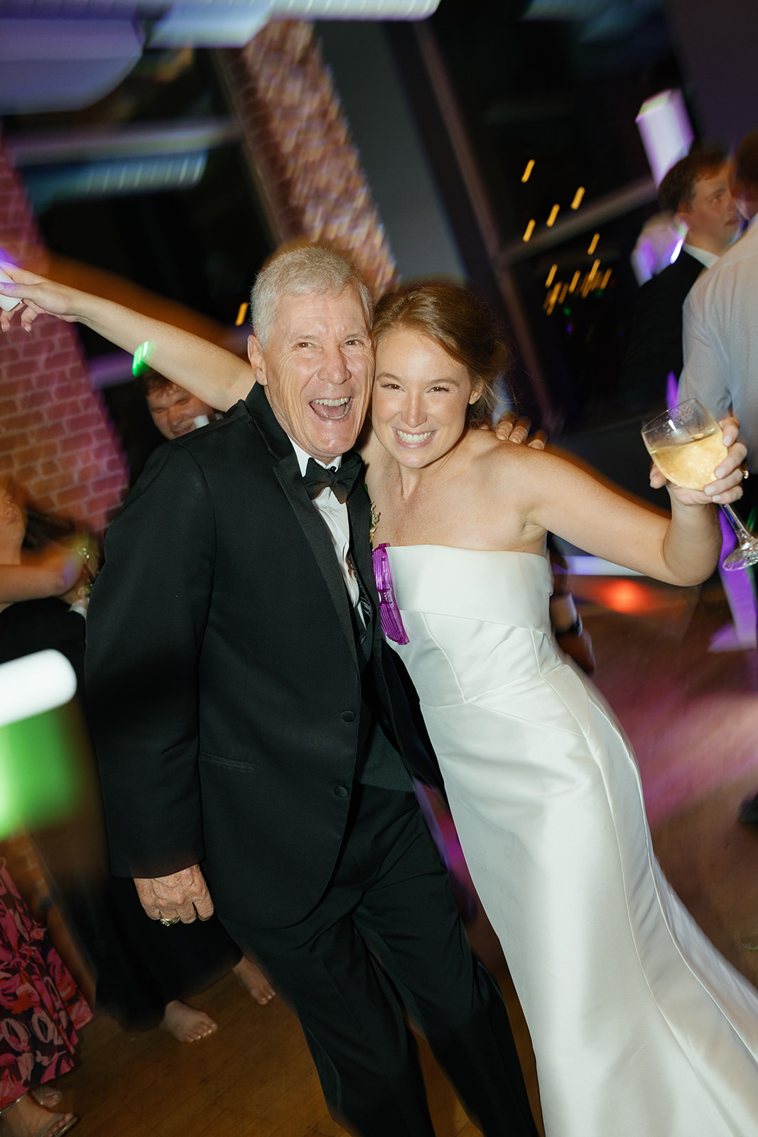 Bride dances and smiles with her dad in a tuxedo while holding a glass of champagne on the reception dance floor.