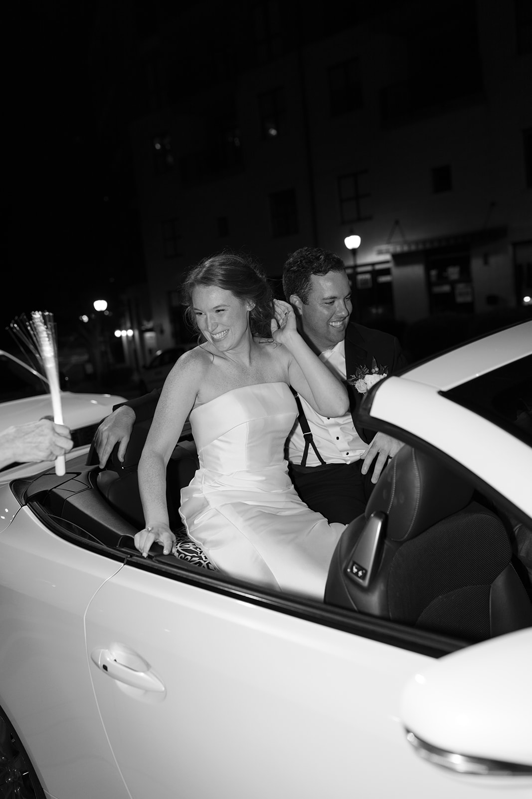 Newlyweds riding away in a white convertible at night, smiling and waving glow wands during their wedding sendoff.