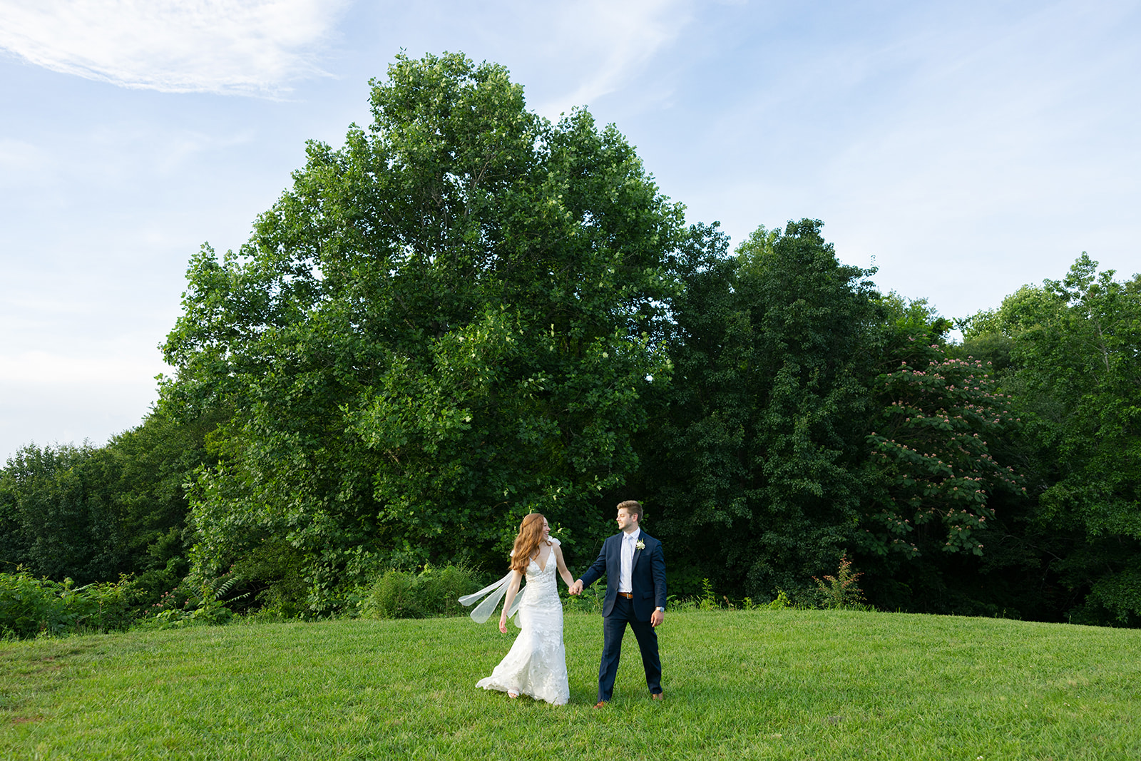 Wide shot of the bride and groom holding hands in a grassy field with tall green trees in the background.