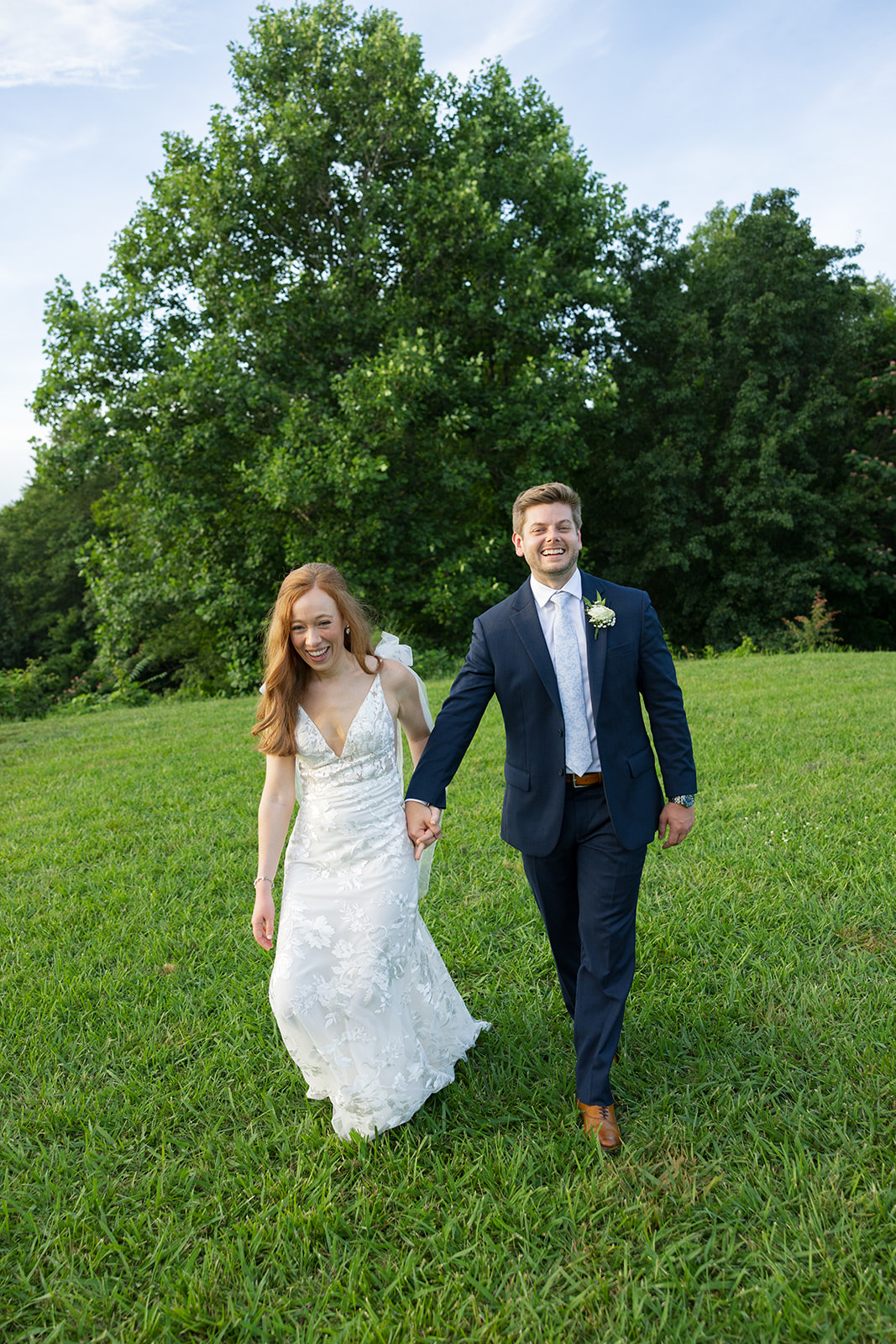 Bride and groom laughing and holding hands while walking together across a sunny green field.