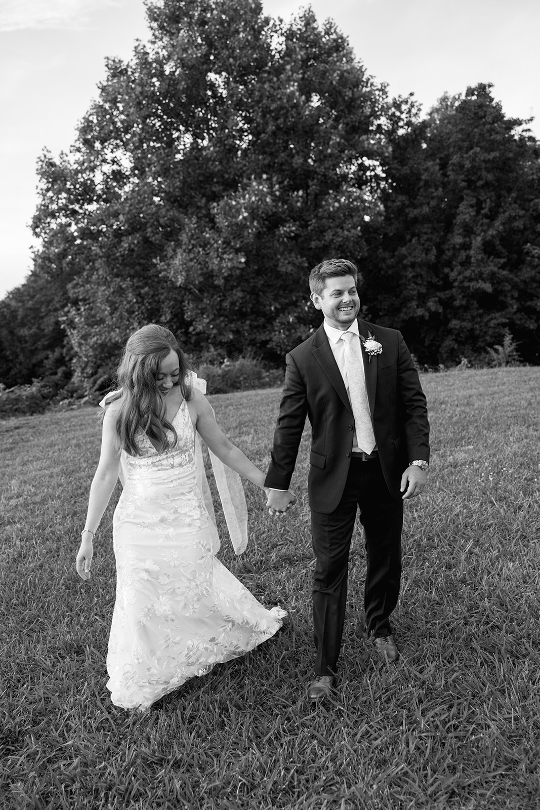 Bride and groom holding hands and walking through a grassy field, smiling together in a candid black-and-white photo shot by a wedding photographer in Greenville, SC