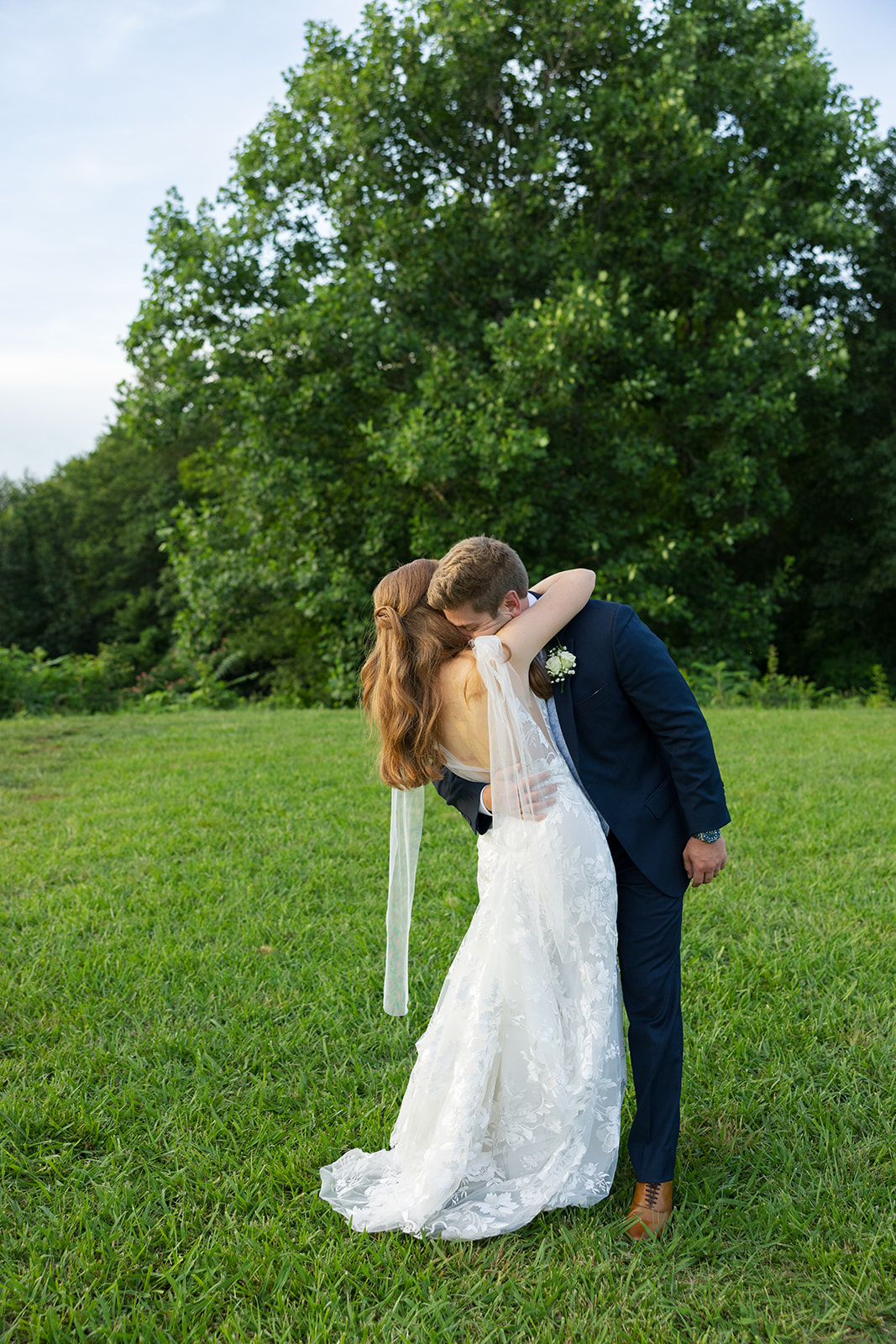 The bride and groom share a romantic kiss in a grassy field with tall green trees in the background.