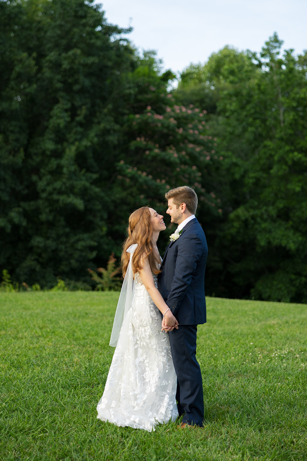 The bride and groom face each other in a grassy field, smiling while holding hands against a backdrop of trees.