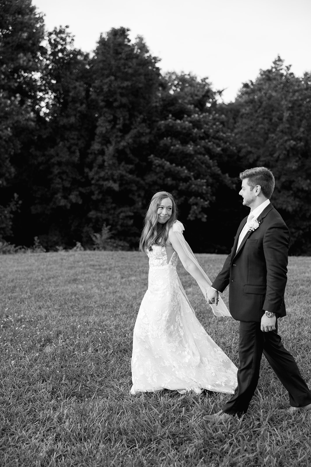 Black and white photo of the couple holding hands and walking through a grassy field, the bride smiling back at the groom.