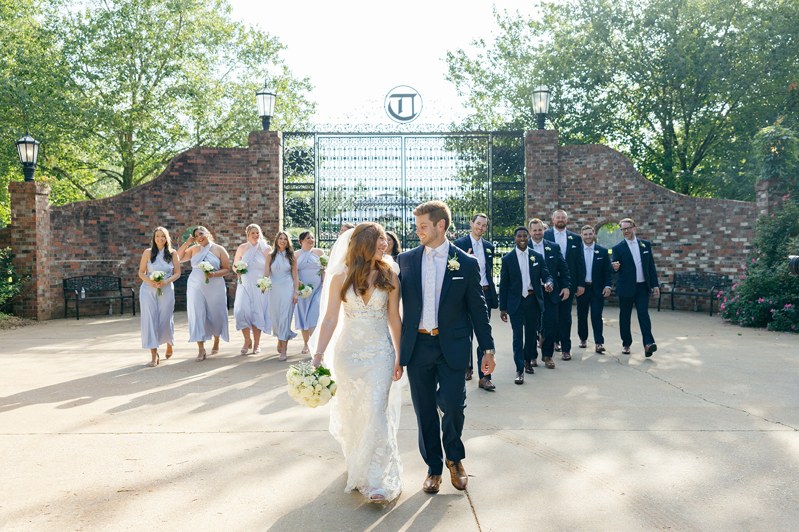 Bride and groom walk hand-in-hand, smiling at each other, with bridesmaids and groomsmen following behind outside a brick gate.