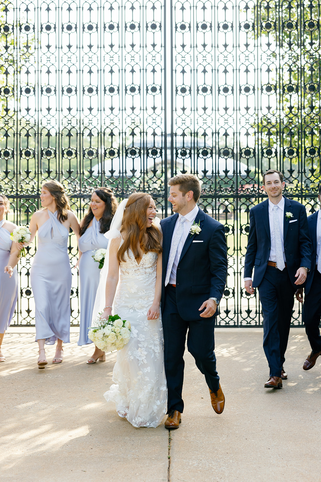 The bride and groom laugh together while walking hand-in-hand, with bridesmaids and groomsmen smiling behind them near an ornate iron gate.