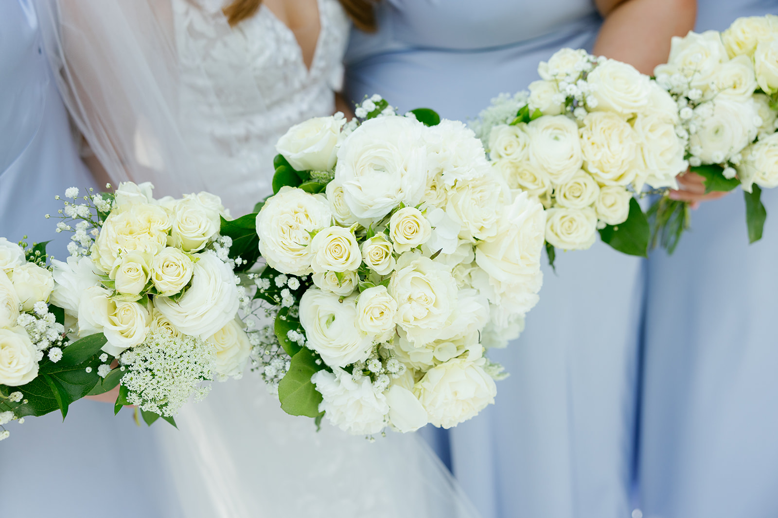 Close-up of the bride and bridesmaids holding bouquets of white roses and greenery, with the women dressed in light blue dresses.