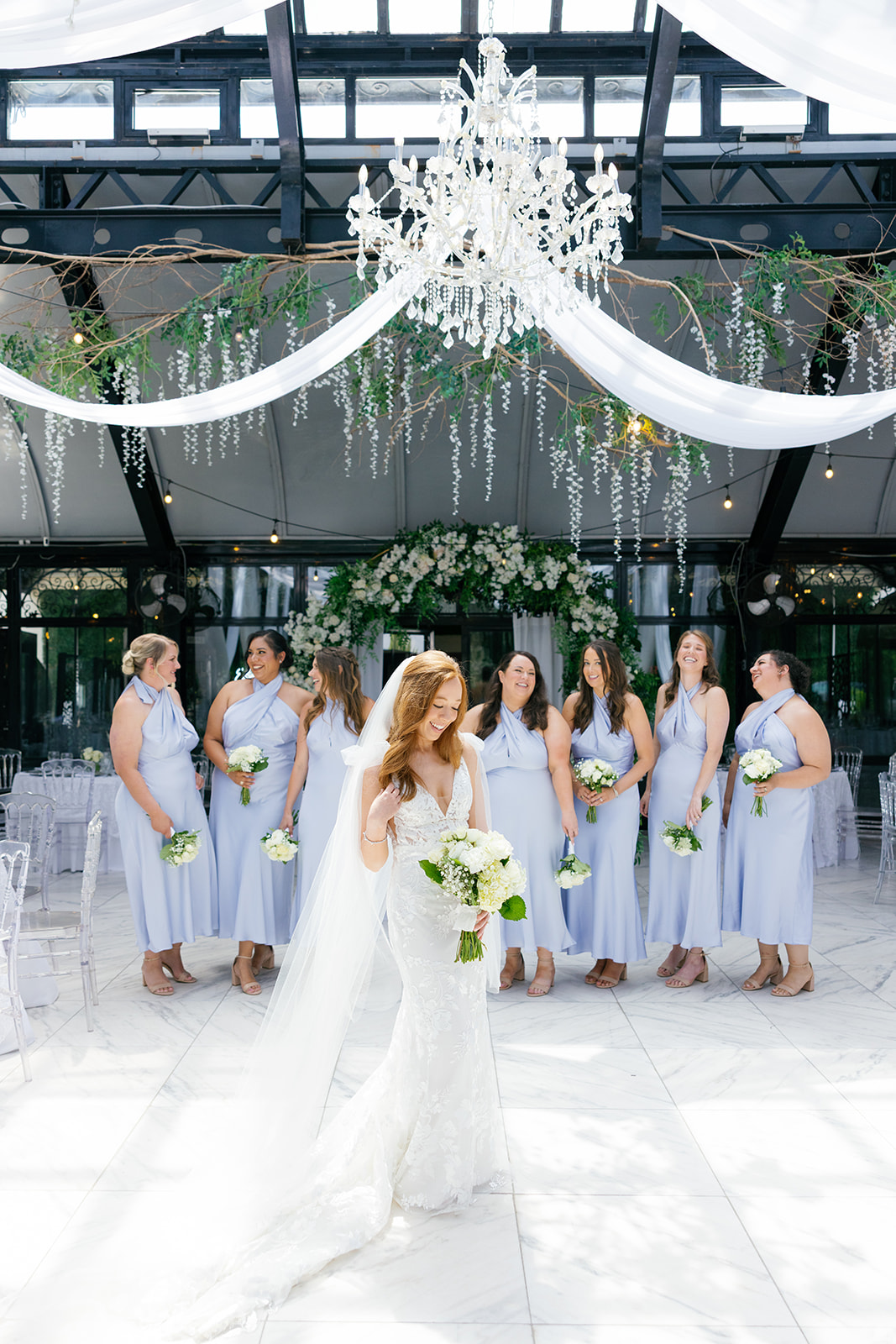Bride stands in front of her bridesmaids inside the reception space, with white draping and a chandelier above.