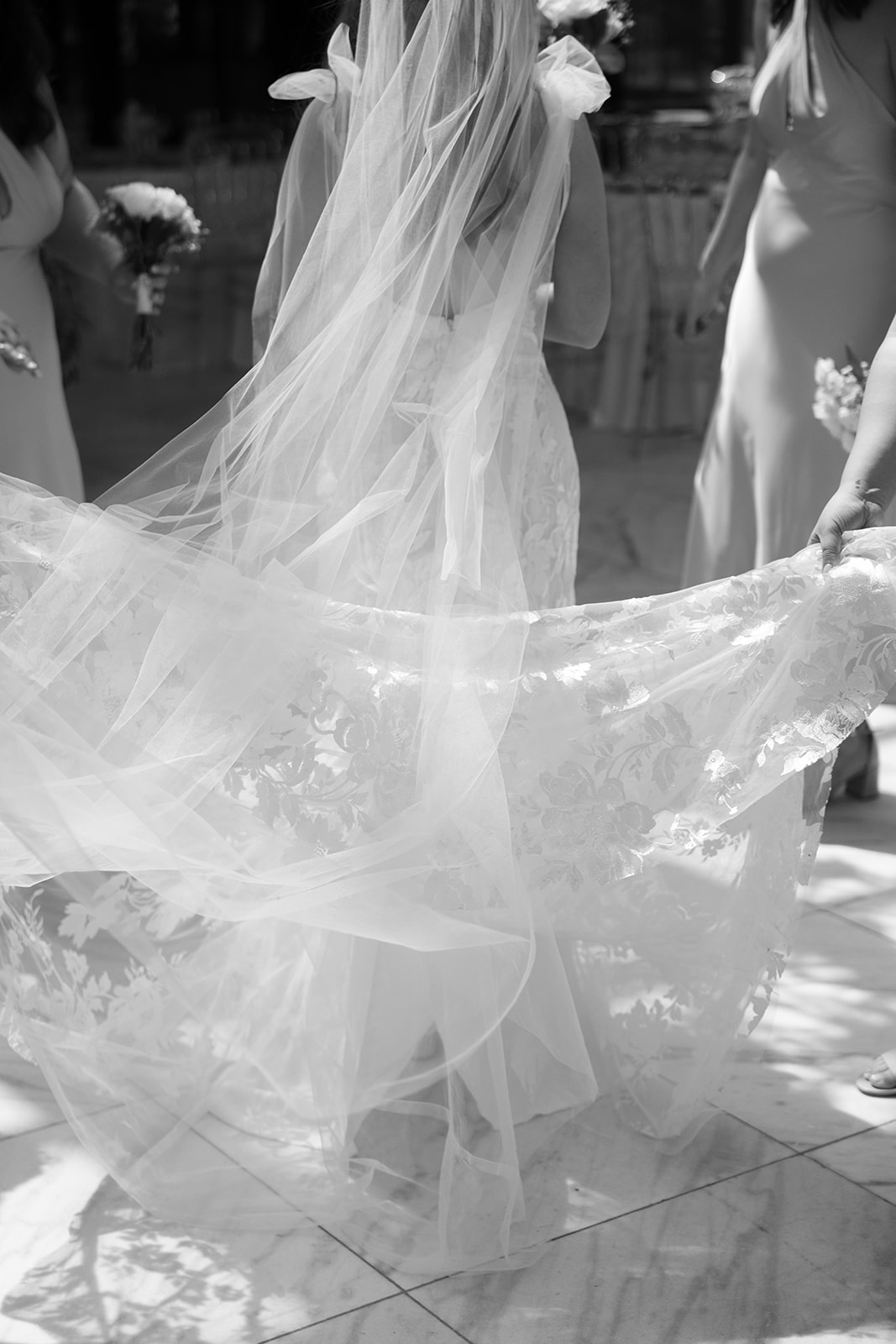 A black and white shot of the bride’s lace gown and long veil being carried as she walks with her bridesmaids.