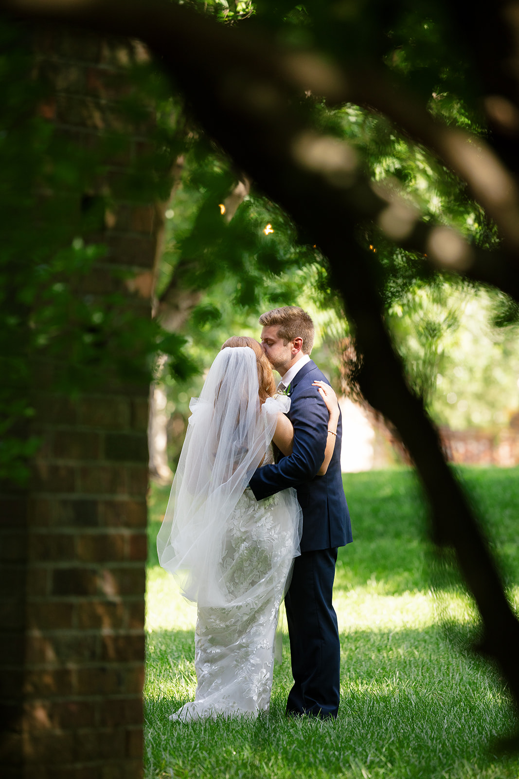 Bride and groom share a quiet kiss on the lawn, framed by trees and soft sunlight.