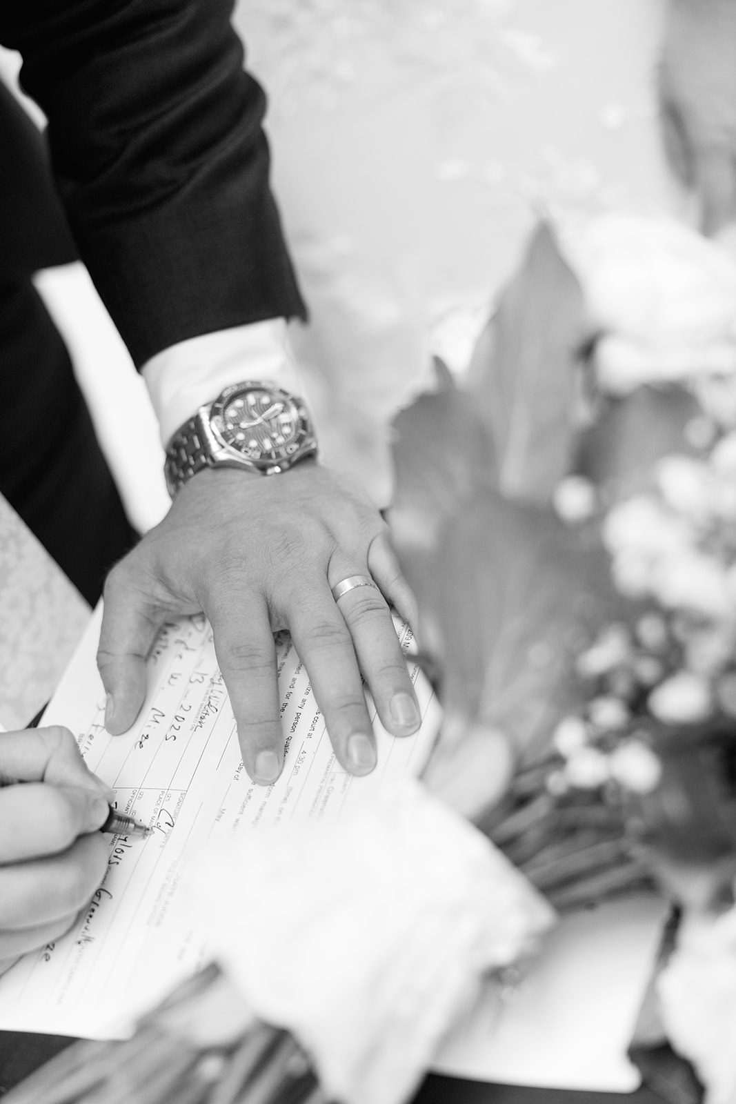 Black and white close-up of the groom’s hand resting on paperwork as the bride signs their marriage license.