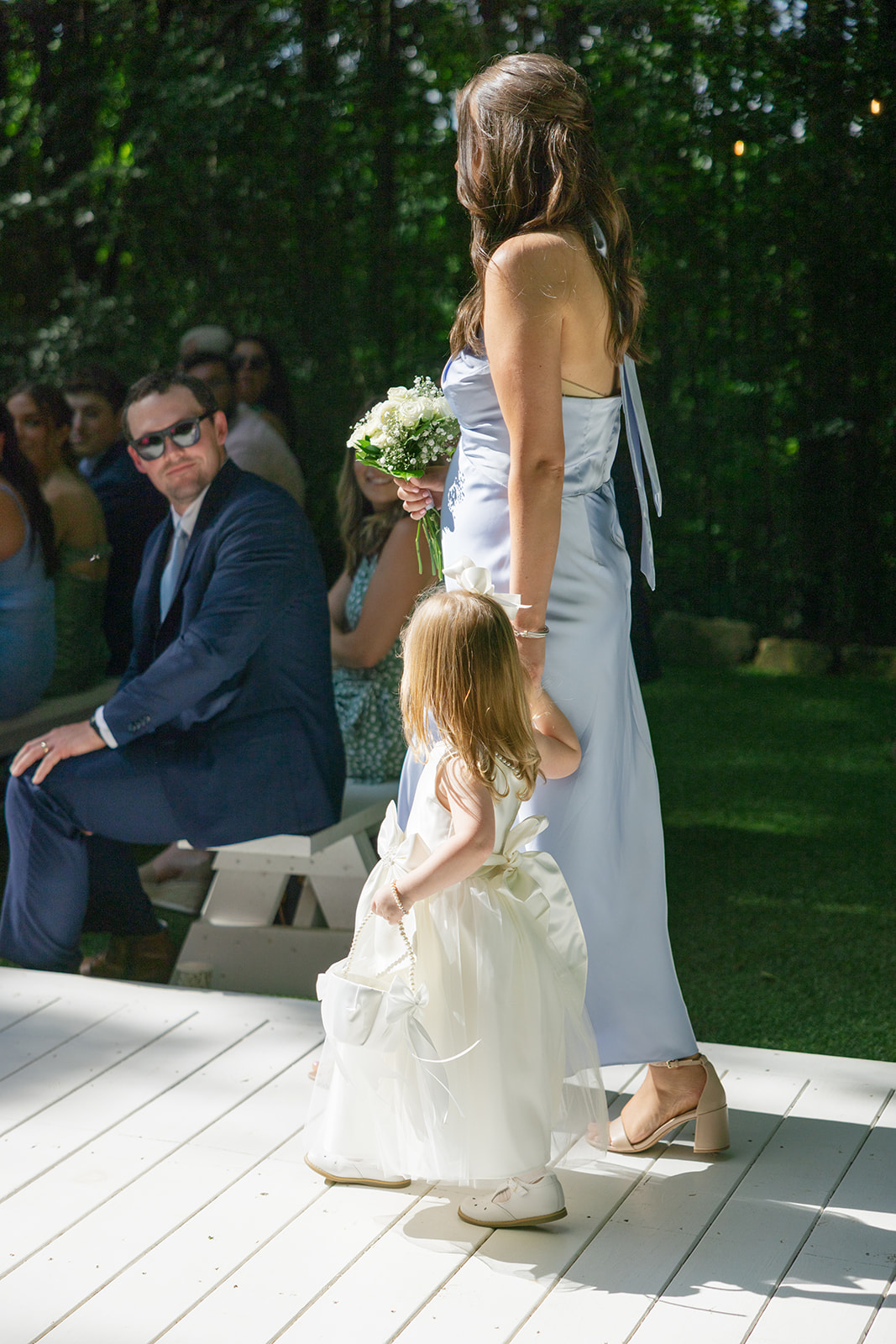 A candid wedding photography moment of a Greenville wedding photographer capturing a bridesmaid in a satin gown holding hands with a flower girl carrying a basket during the ceremony at Edinburgh West.
