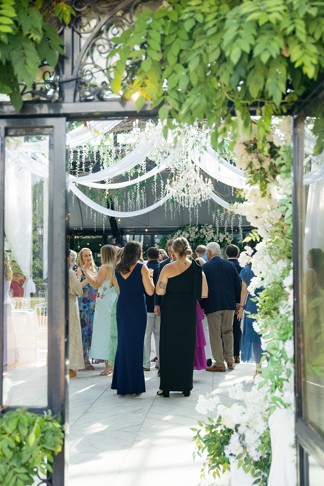 A view through floral-adorned doors showing wedding guests gathered under white draping and chandeliers.