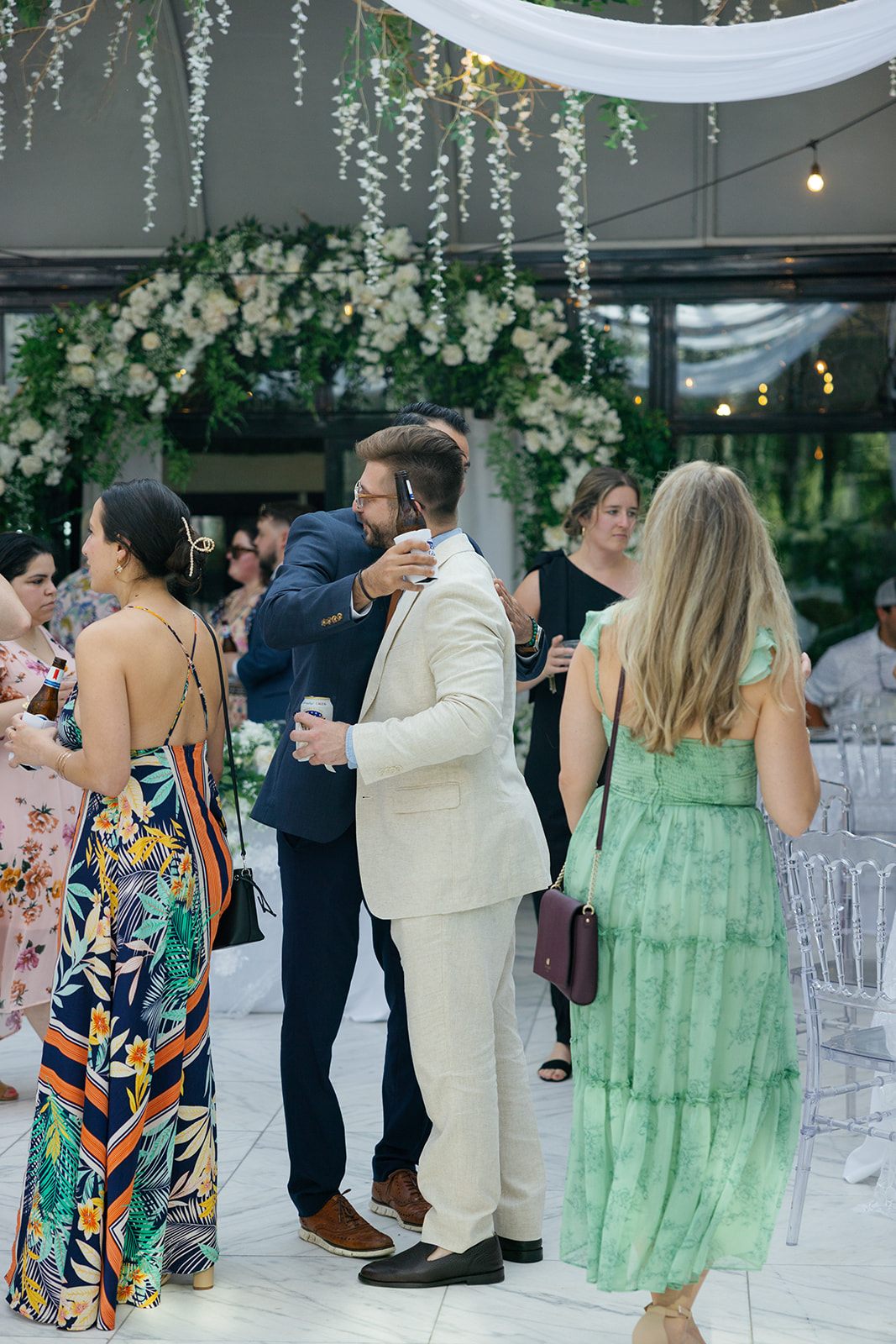 A guest in a cream suit hugs another in a navy suit while holding a beer, surrounded by mingling guests.